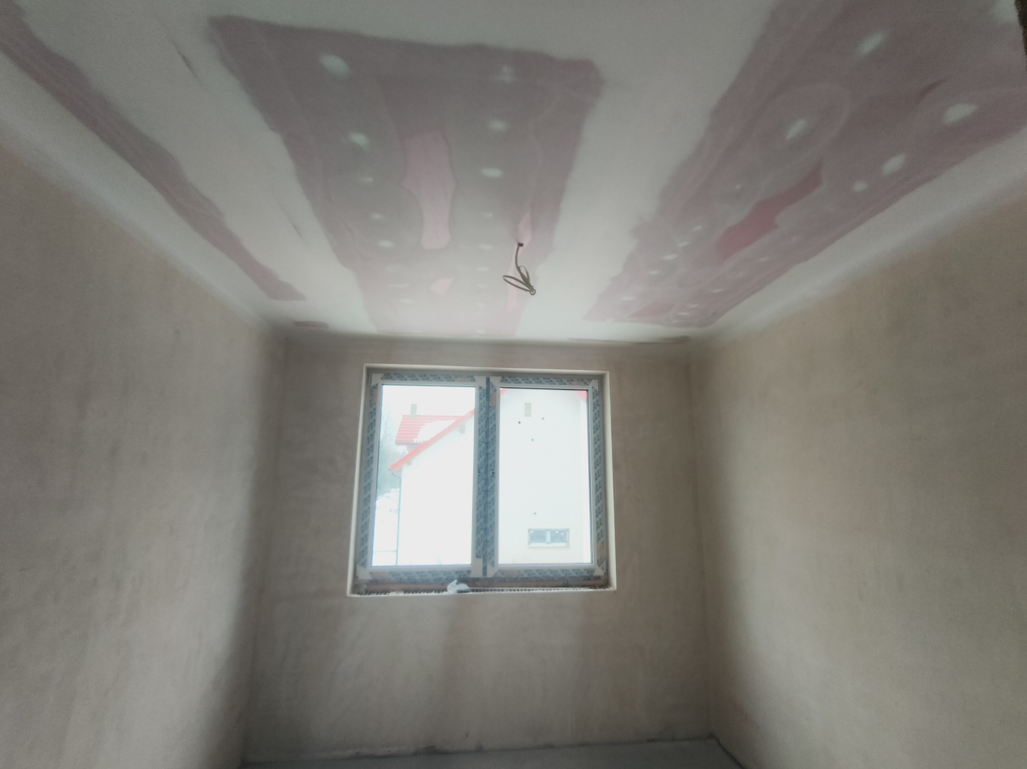 Pokój w trakcie szpachlowania: sufit z widocznymi łatami szpachli, ściany przygotowane do malowania, okno z widokiem na dom. Wnętrze w stanie surowym.