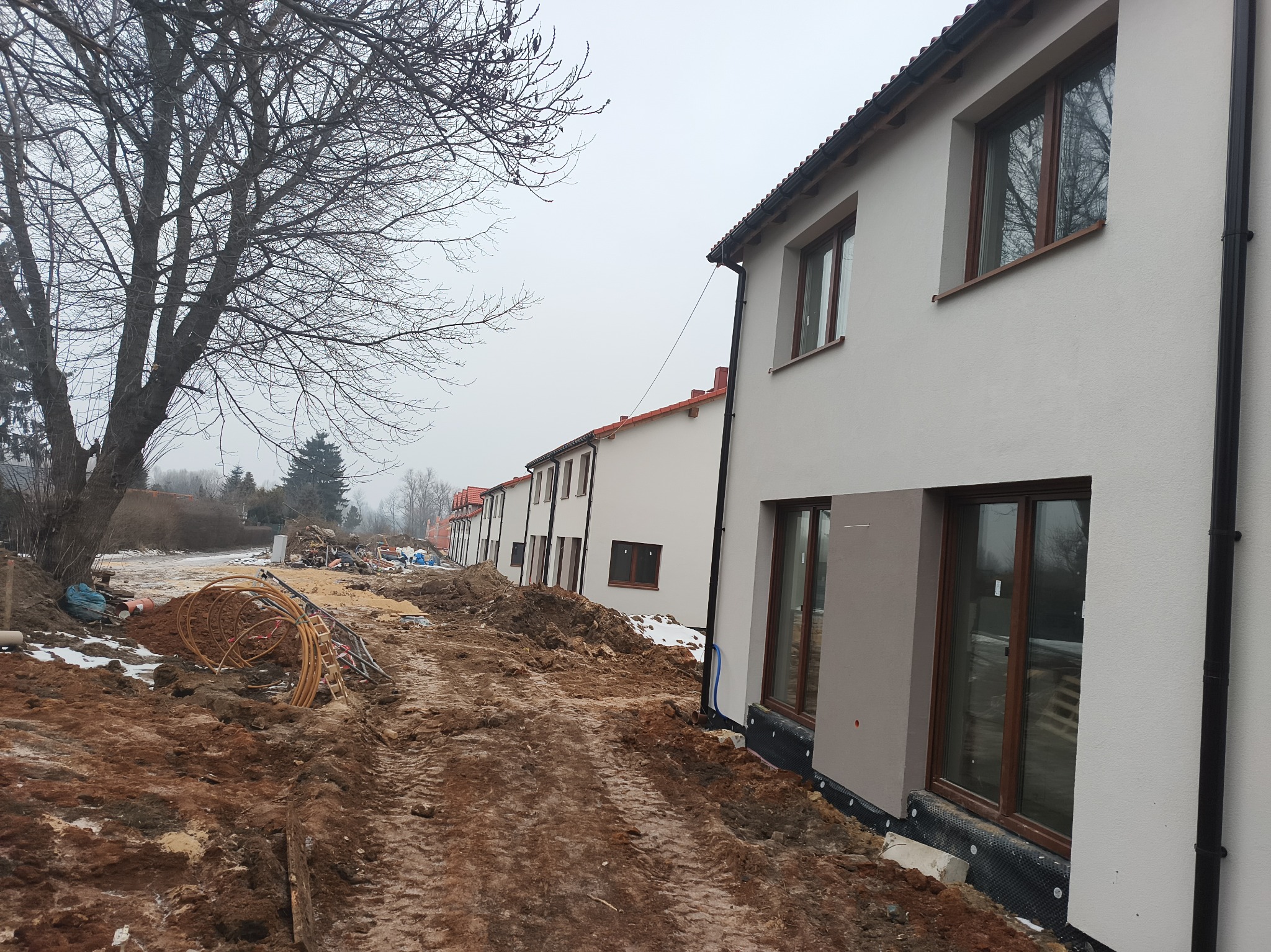Nowe budynki w trakcie wykończenia, widoczne okna z brązowymi ramami, świeży tynk i przygotowany teren wokół. Prace ziemne w toku, widoczne materiały budowlane.
