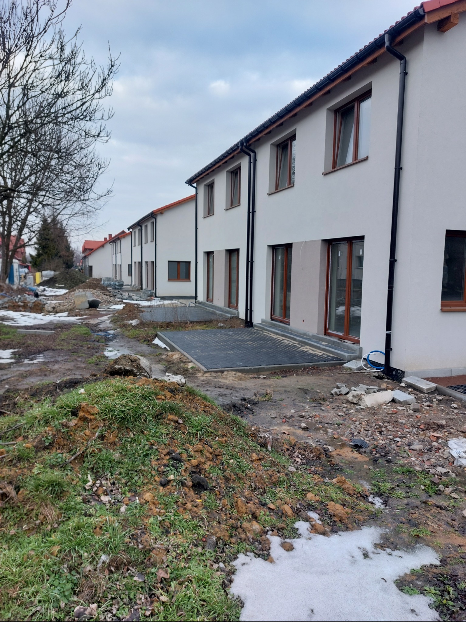 Szereg identycznych, jasnych budynków z brązowymi oknami i czarnymi rynnami. Ułożona kostka brukowa przed wejściami. Teren wokół w trakcie prac ziemnych.