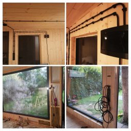 GK Gabriel Kukułowicz - Wnętrze drewnianego domku z widoczną instalacją elektryczną natynkową w czarnych peszlach, duże okno z widokiem na zewnątrz, etap prac wykończeniowych.
