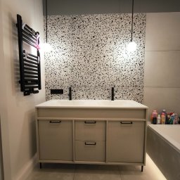 GK Gabriel Kukułowicz - Beżowa szafka łazienkowa z czarnymi uchwytami i umywalką, na tle ściany z mozaiką i grzejnika drabinkowego. Minimalistyczne lampy wiszące.