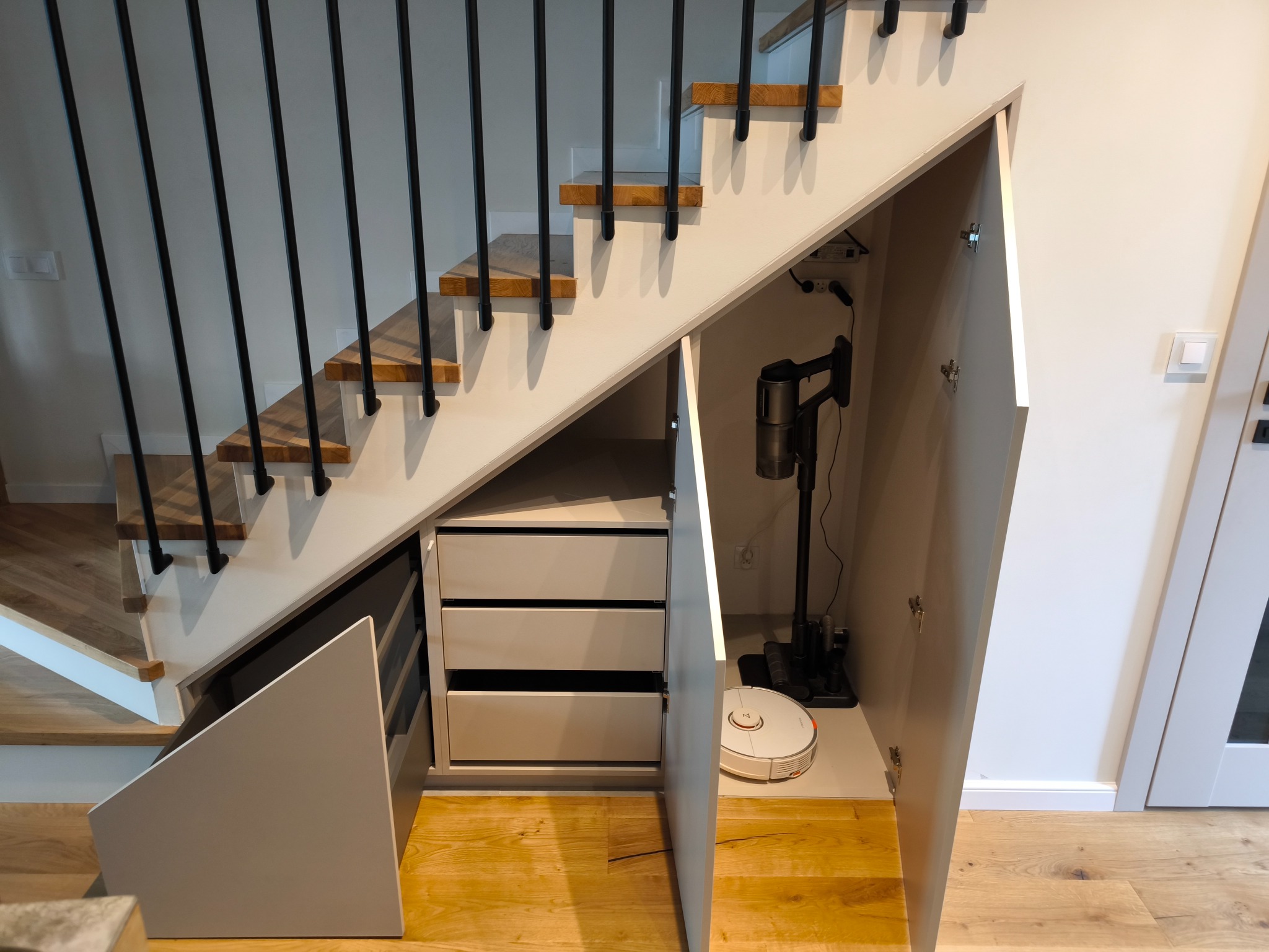 Szafka pod schodami z otwartymi drzwiczkami, wewnątrz odkurzacz pionowy i robot sprzątający. Widoczne drewniane schody z czarną balustradą i podłoga z paneli.