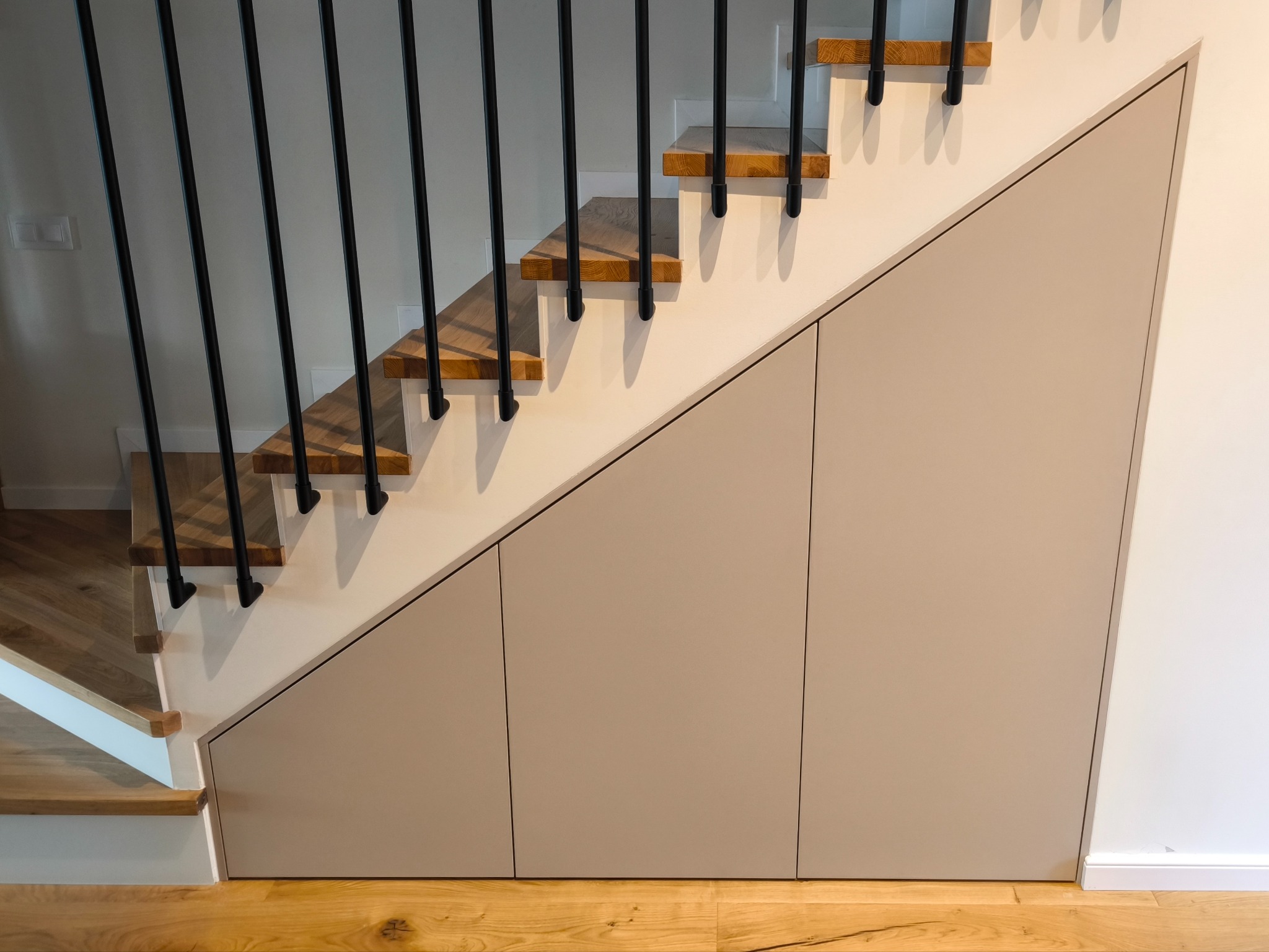 Schowek pod schodami z drewnianymi stopniami i czarną balustradą. Beżowe fronty szafek wkomponowane w przestrzeń pod schodami. Parkiet na podłodze.