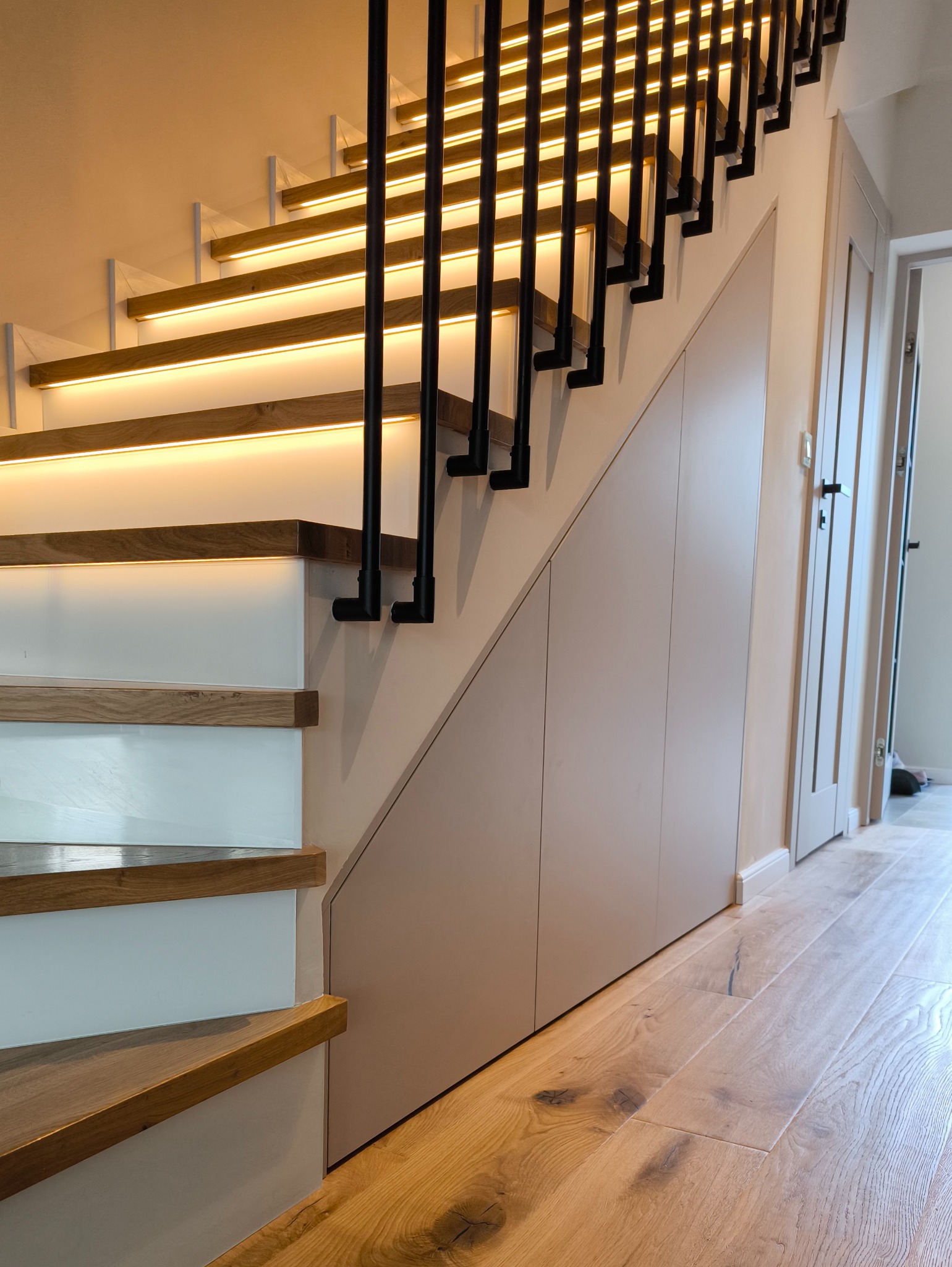 Nowoczesne schody z podświetleniem LED i czarną balustradą, zabudowane szafkami w kolorze beżowym, parkiet dębowy. Minimalistyczny design.