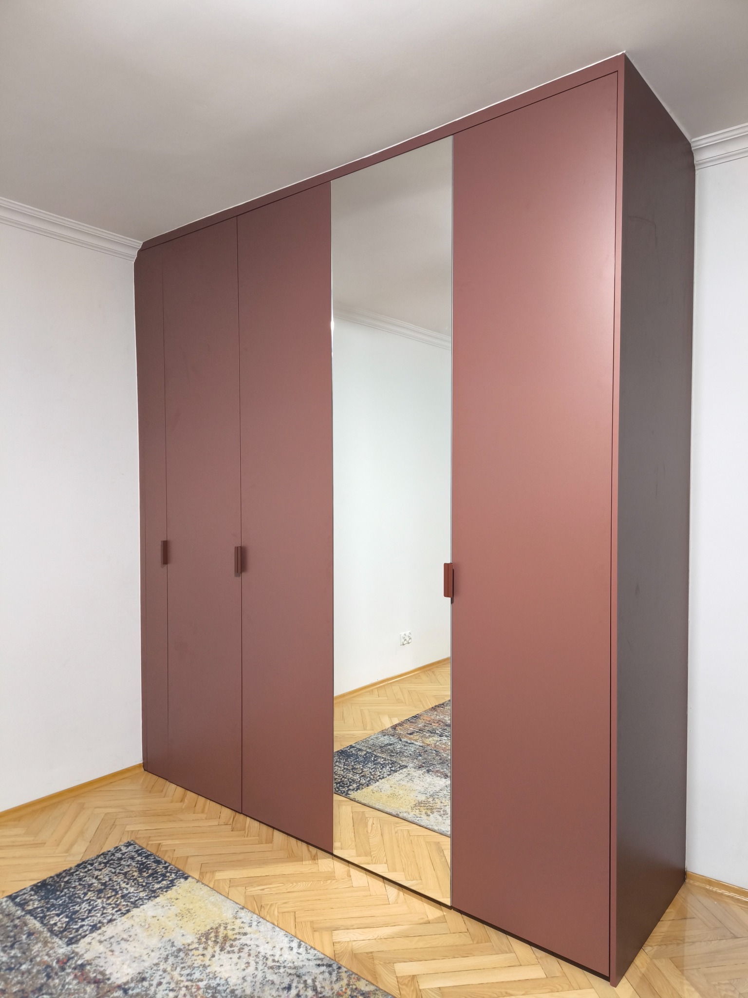 Wysoka, matowa szafa wnękowa w odcieniach terakoty z lustrzanym frontem, odbijającym wzorzysty dywan na parkiecie. Minimalistyczne uchwyty, białe ściany i sufit z dekoracyjną listwą.