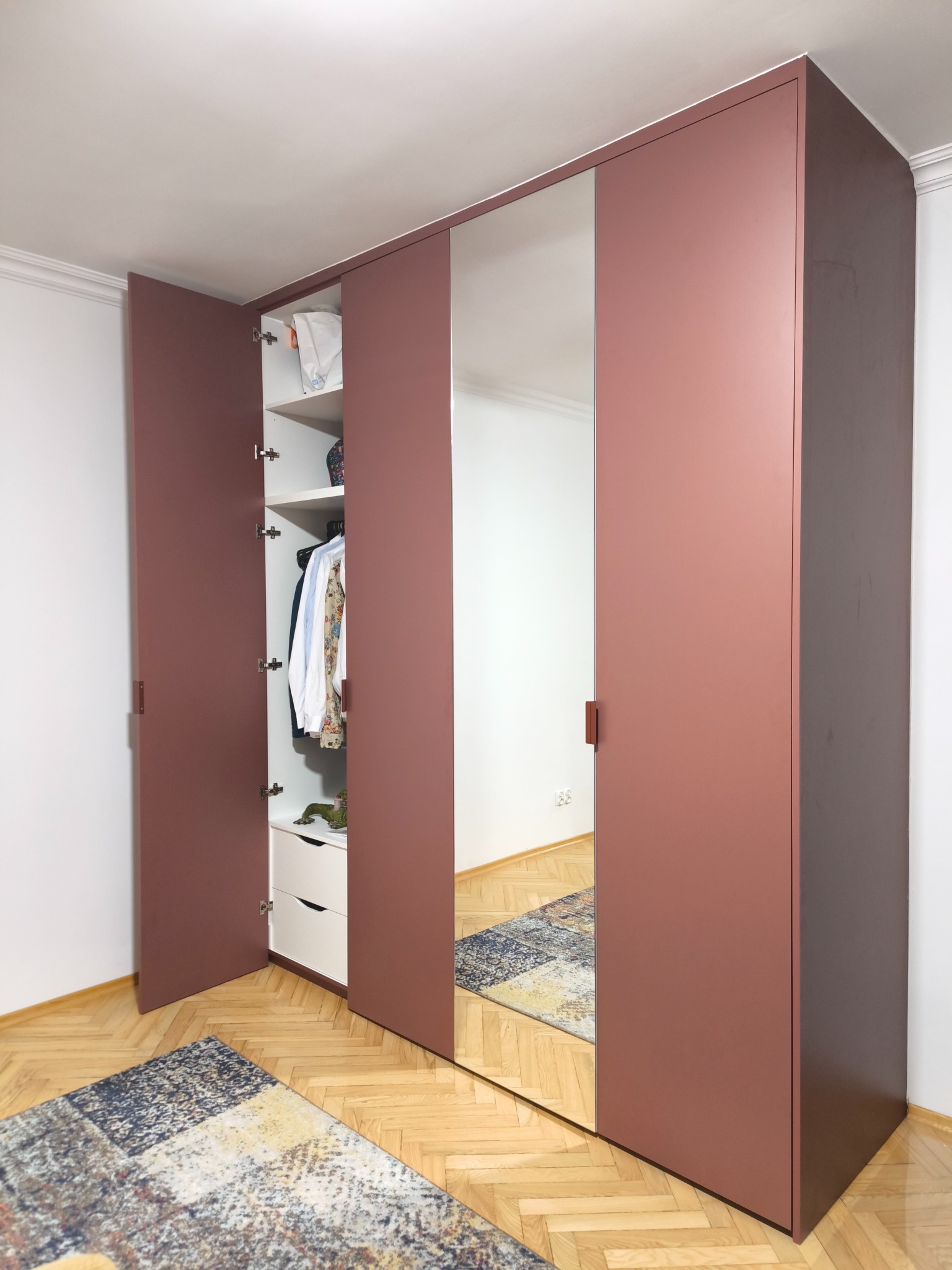 Wnętrze: Szafa wnękowa z lustrem i otwartymi drzwiami, ukazująca ubrania na półkach. Kolorystyka: odcienie różu i bieli. Parkiet w jodełkę, dywan w tle.
