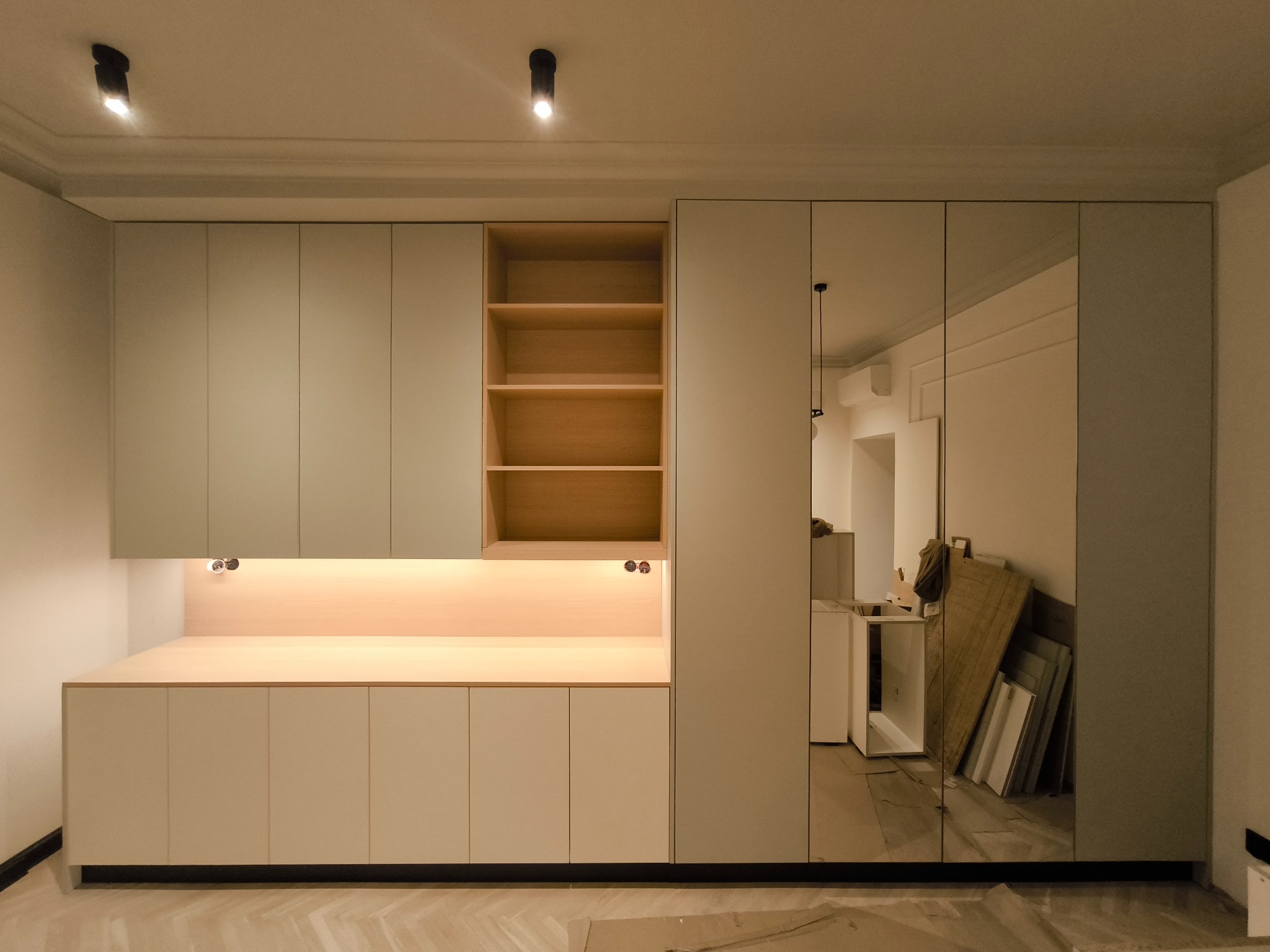 Jasna zabudowa meblowa z szafkami, półkami i lustrzanymi drzwiami, oświetlona punktowo, z widocznym parkietem i elementami wykończeniowymi w tle.