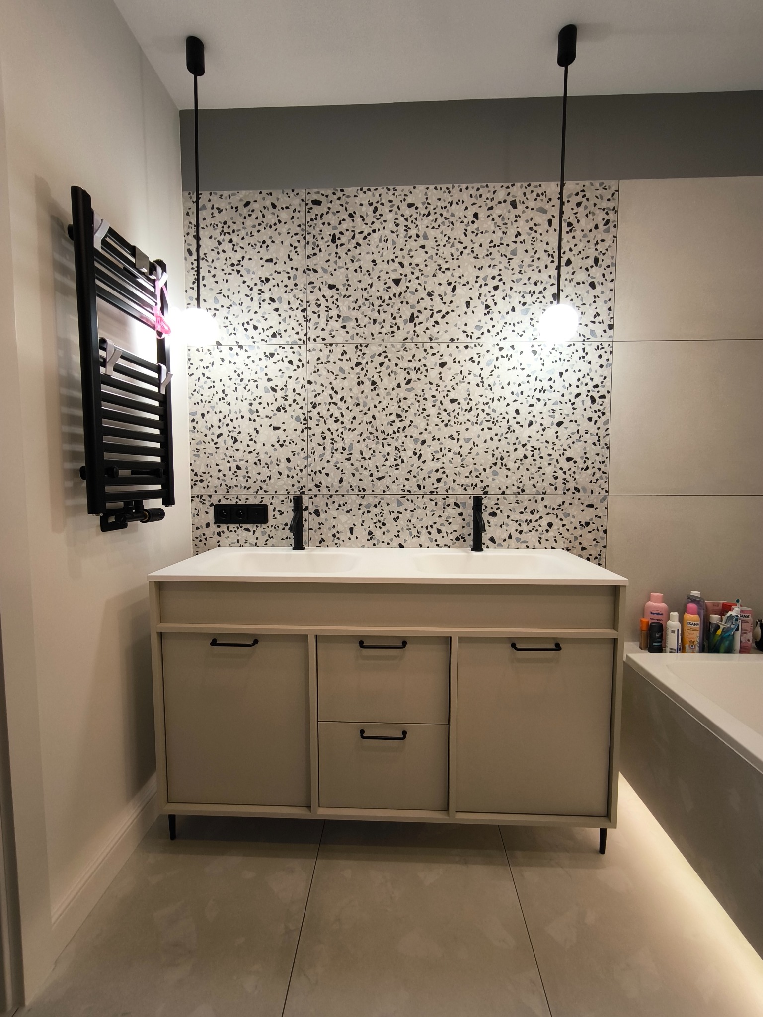 Beżowa szafka łazienkowa z czarnymi uchwytami i umywalką, na tle ściany z mozaiką i grzejnika drabinkowego. Minimalistyczne lampy wiszące.