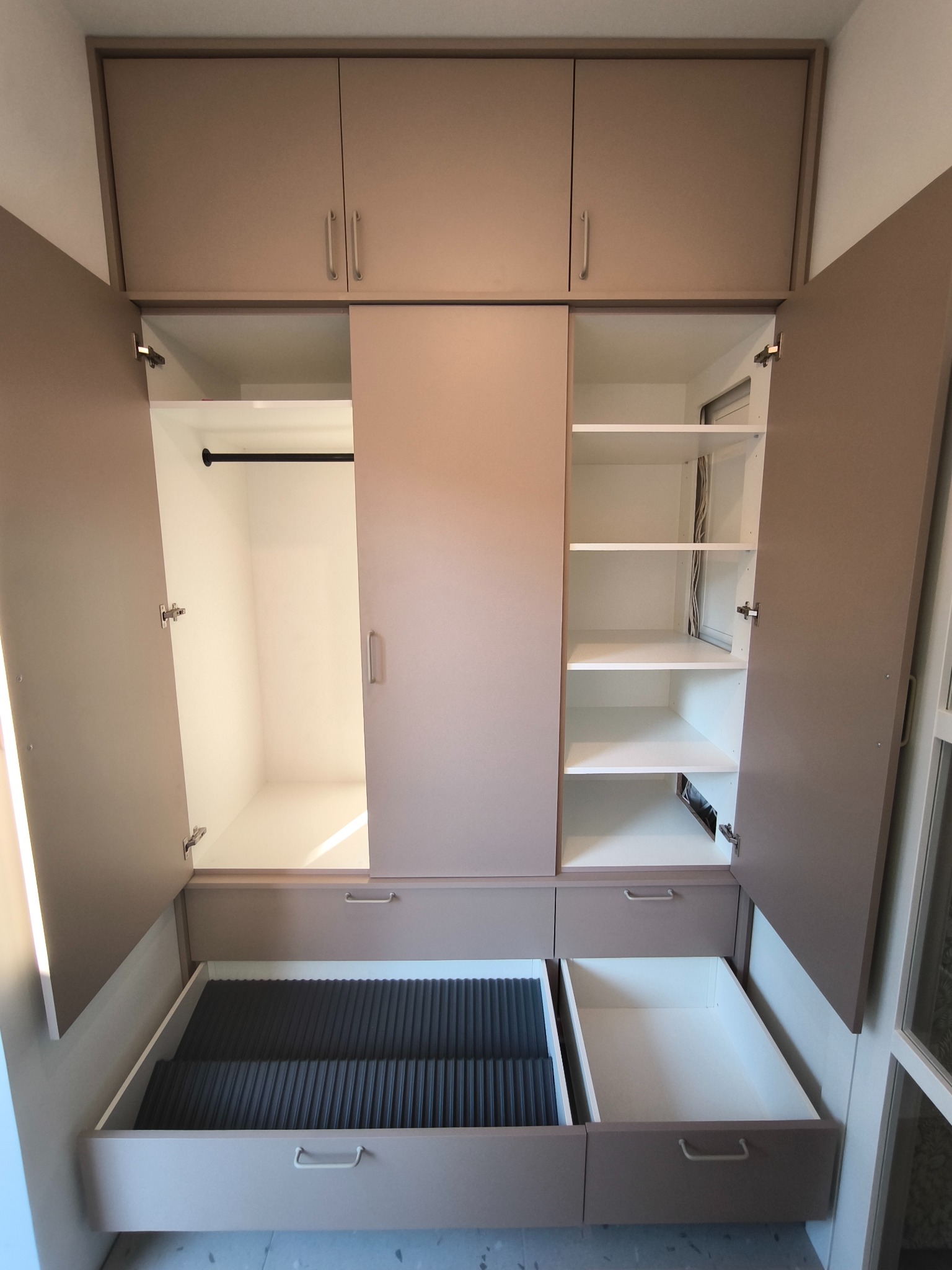 Beżowa szafa wnękowa z otwartymi drzwiami i szufladami, widoczne półki i drążek na ubrania. Minimalistyczny design, jasne wnętrze, szuflady z różnym wyposażeniem.