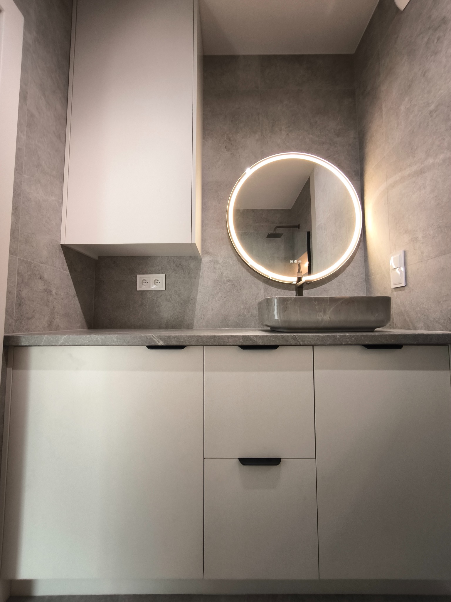 Nowoczesna łazienka z szafką pod umywalkę w kolorze złamanej bieli, okrągłym lustrem z podświetleniem LED i szarą umywalką nablatową. Minimalistyczny design.