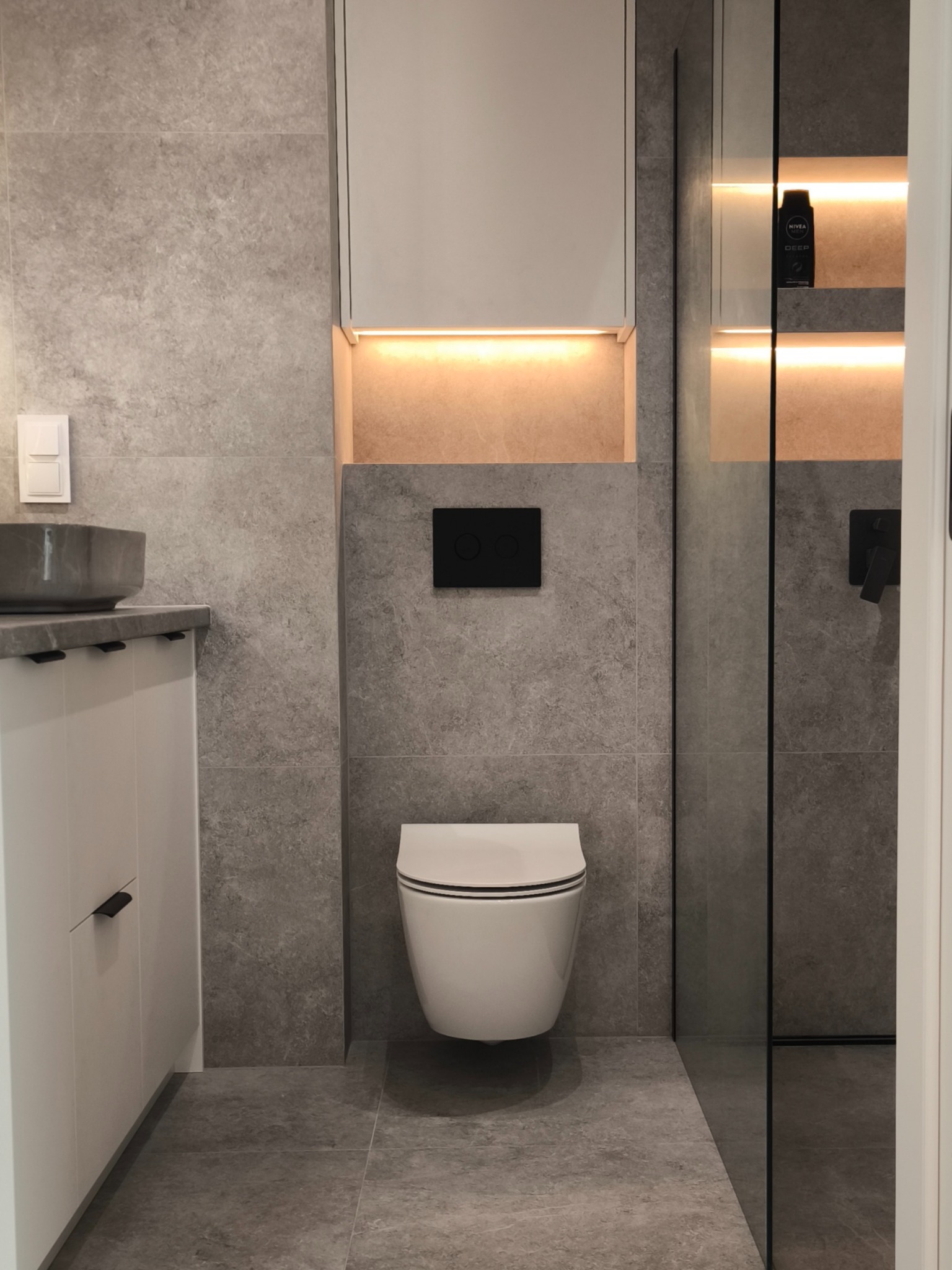 Nowoczesna łazienka z szarą armaturą i podświetlanymi niszami. Umywalka nablatowa, wisząca toaleta i kabina prysznicowa ze szklaną ścianką.