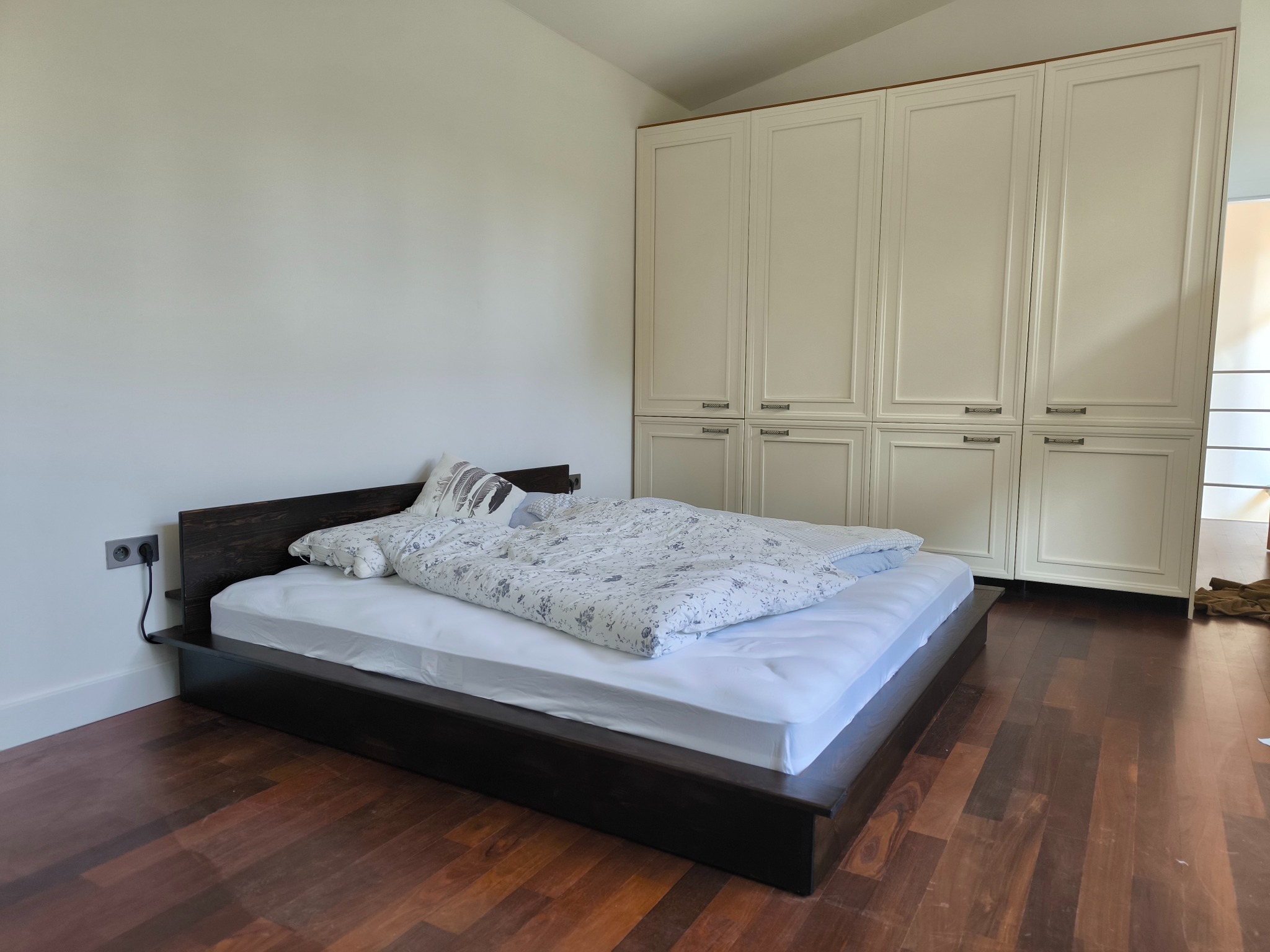 Nowoczesna sypialnia z niskim łóżkiem z ciemnego drewna i białą pościelą. W tle biała szafa w zabudowie z frezowanymi frontami i metalowymi uchwytami.