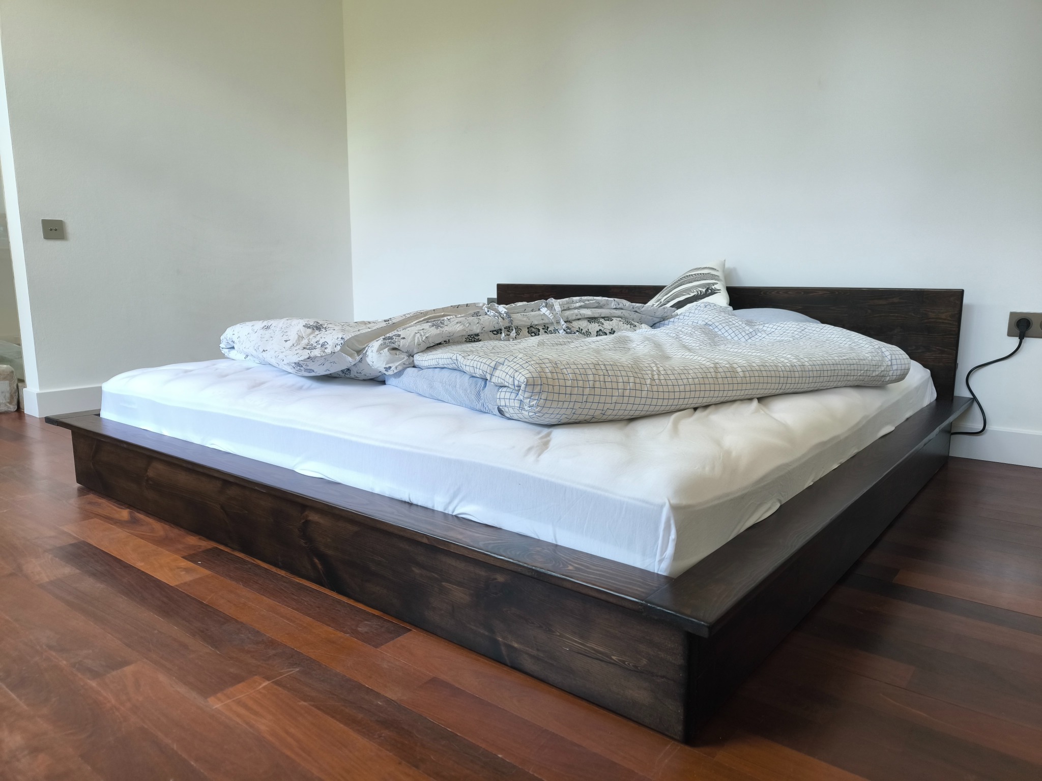 Drewniane łóżko na niskiej ramie z ciemnego drewna, z pościelą w odcieniach bieli i szarości, na tle białej ściany i drewnianej podłogi. Minimalistyczny design.