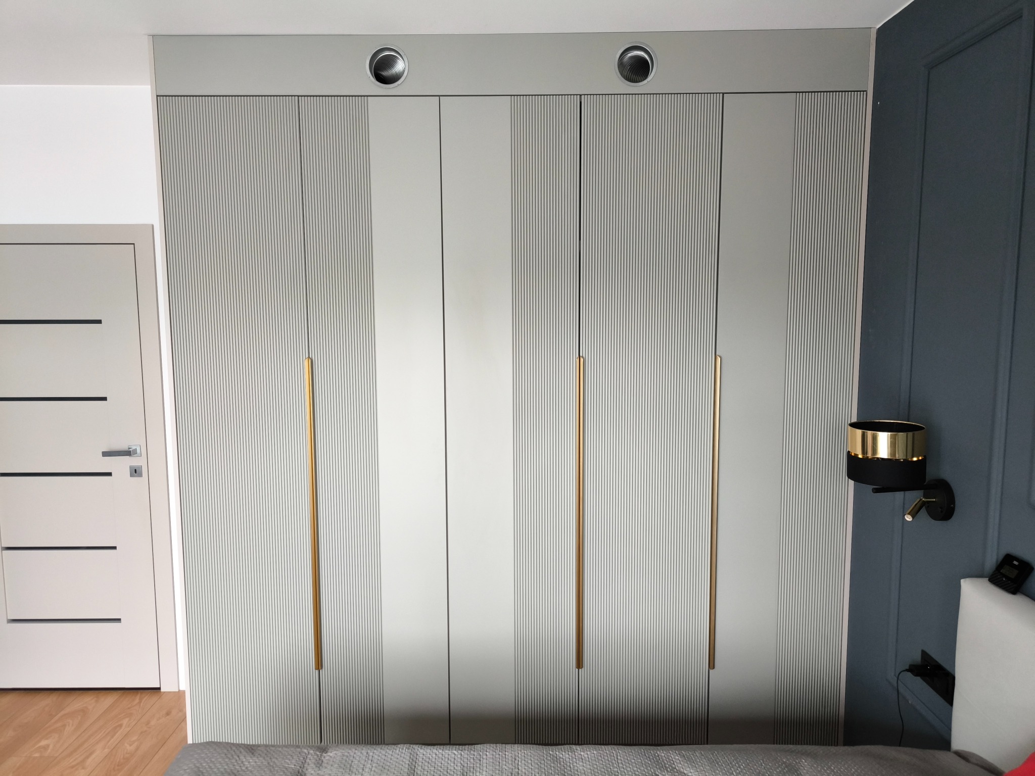 Szara szafa wnękowa z ryflowanymi frontami i złotymi uchwytami, wkomponowana w aranżację sypialni z ciemną ścianą i lampką nocną. Widoczne detale wykończenia.