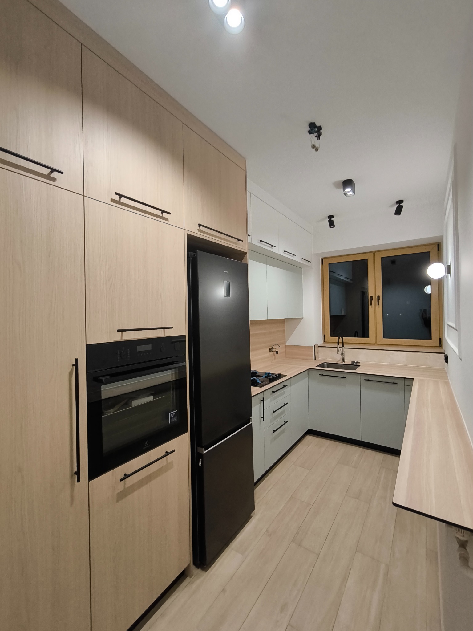 Nowoczesna kuchnia z drewnianymi frontami szafek, czarnym sprzętem AGD i szarymi dolnymi szafkami. Uwagę przykuwają podłużne, czarne uchwyty i oświetlenie punktowe.