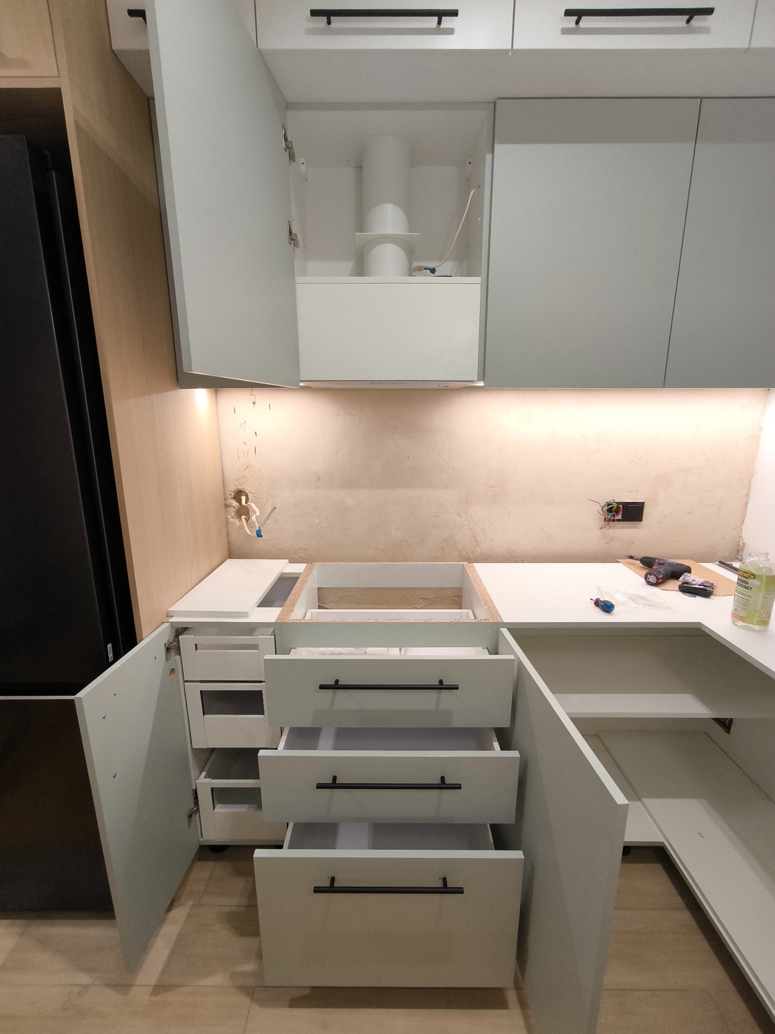Montaż szuflad w jasnej kuchni na wymiar z czarnymi uchwytami. Widoczne narzędzia i niedokończone podłączenia elektryczne. Oświetlenie LED pod szafkami.