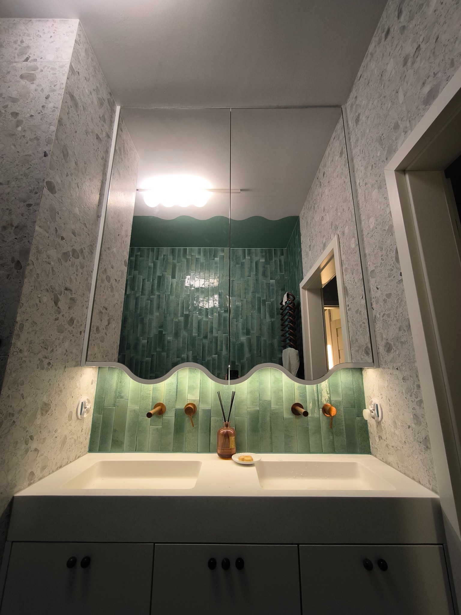 Nowoczesna łazienka z szafką pod umywalką, zielonymi płytkami i lustrem. Umywalka z dwoma kranami w kolorze miedzi. Ściany wykończone płytkami imitującymi lastryko.