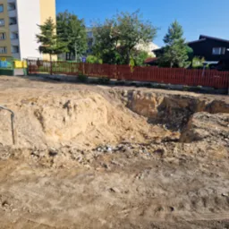 Wykonany wykop w ziemi na terenie budowy, widoczne ślady opon i gruz, w tle zabudowa mieszkalna i drewniany płot.