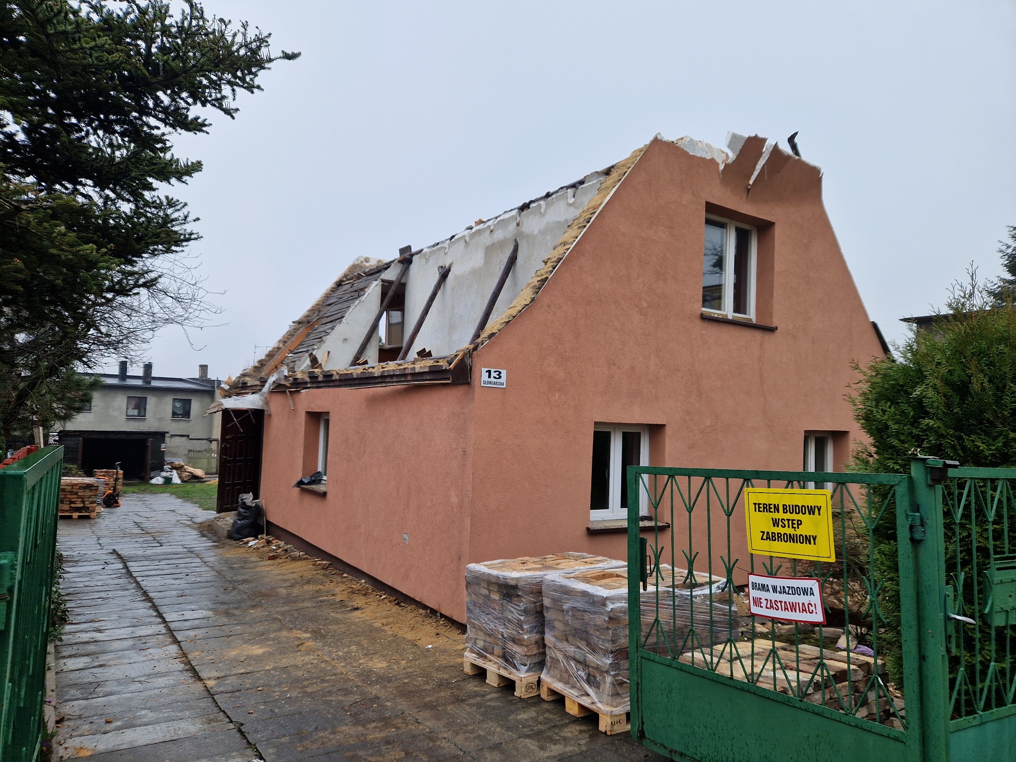 Dom z częściowo zdemontowanym dachem, widoczne elementy konstrukcyjne, przygotowane materiały budowlane na paletach przy ogrodzeniu z tabliczką informacyjną o terenie budowy.
