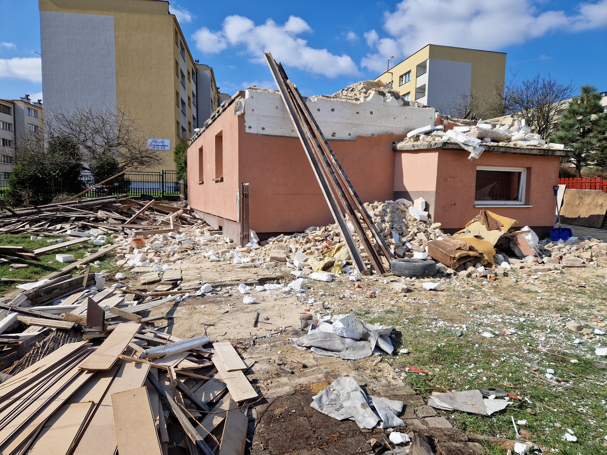 Częściowo rozebrany, jednopiętrowy budynek z odsłoniętą konstrukcją dachu i gruzem na ziemi, z drabiną opartą o ścianę, na tle budynków mieszkalnych i nieba.