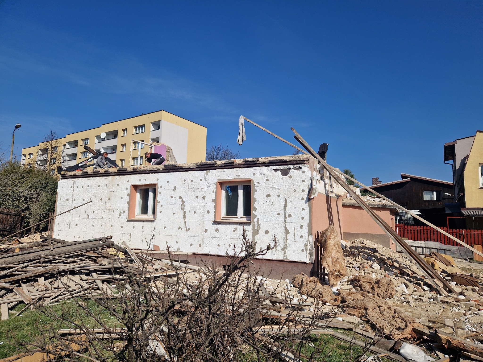 Dwóch pracowników na dachu częściowo rozebranego budynku, widoczne pozostałości konstrukcji, gruz i izolacja, w tle blok mieszkalny i błękitne niebo.