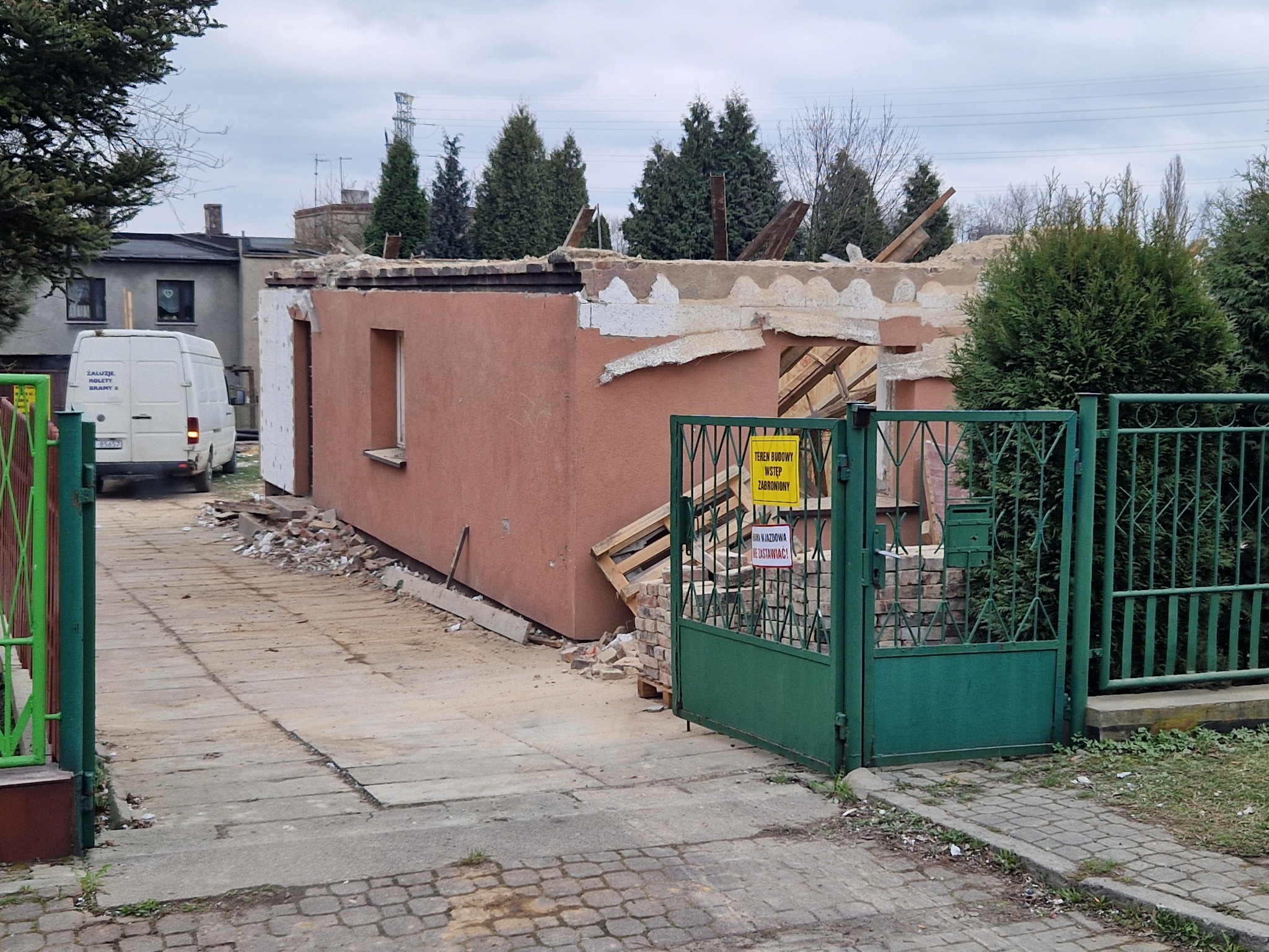 Zdjęcie z zewnątrz przedstawia częściowo rozebrany budynek z cegły, z widocznymi fragmentami dachu i ścian, teren budowy zabezpieczony zielonym ogrodzeniem z tabliczką ostrzegawczą, w tle biały...