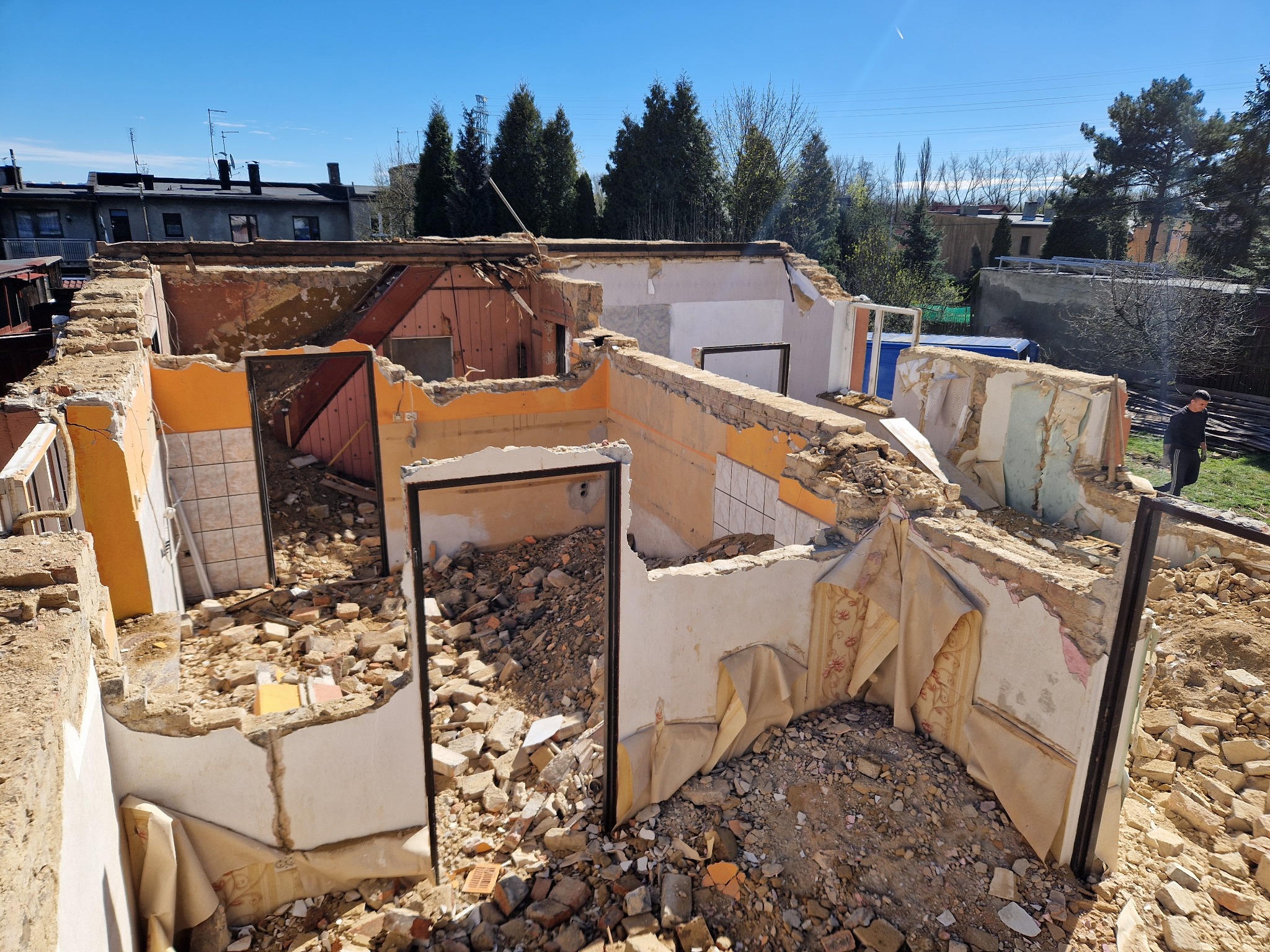 Wnętrze zrujnowanego domu z resztkami tapet i gruzem, widok na zewnątrz, gdzie stoi mężczyzna i widać drzewa.