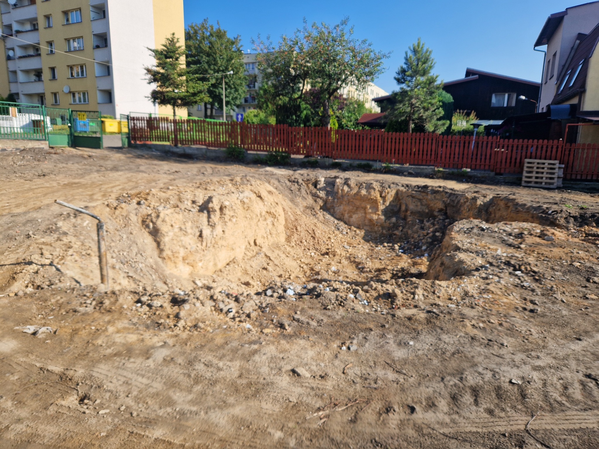 Wykonany wykop w ziemi na terenie budowy, widoczne ślady opon i gruz, w tle zabudowa mieszkalna i drewniany płot.