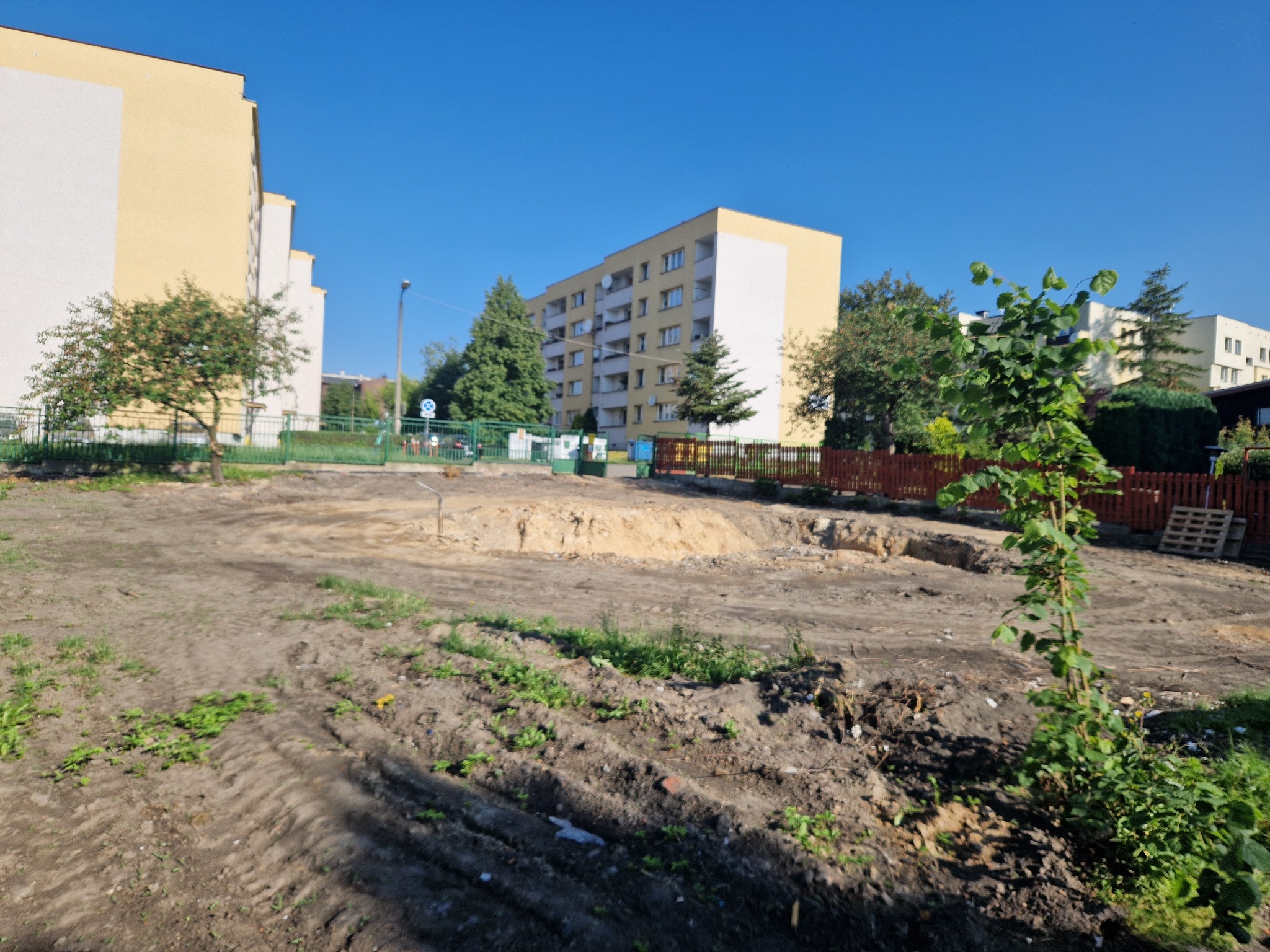 Teren po usunięciu nawierzchni, przygotowany pod budowę, z widocznymi śladami opon, w tle bloki mieszkalne.