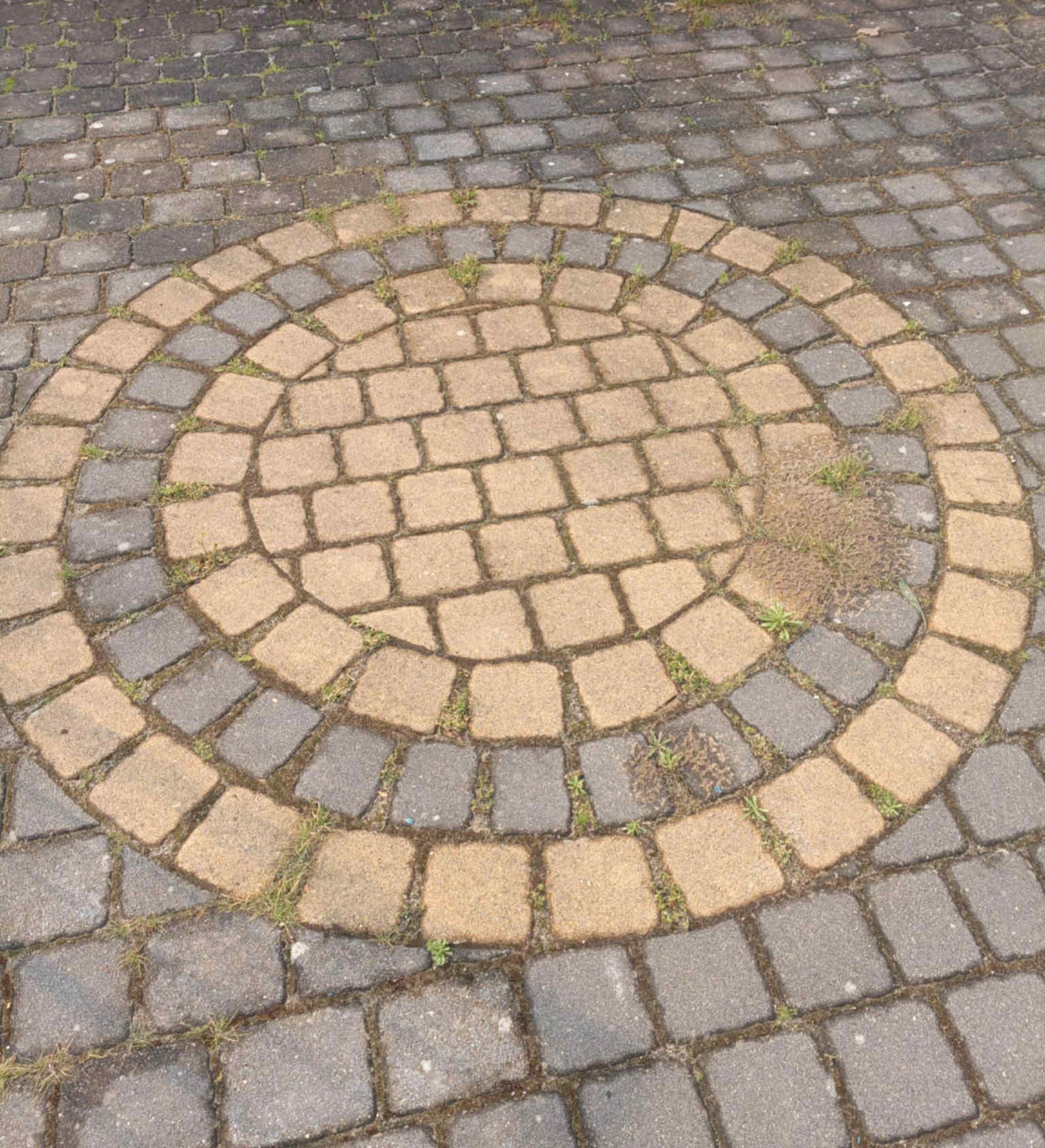 Kostka brukowa ułożona w okrągły wzór, z widocznymi zabrudzeniami i porostami między kostkami. Drobne chwasty wyrastają spomiędzy elementów.