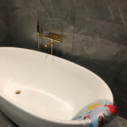 Albert Brzezin - Nowoczesna, wolnostojąca wanna owalna z armaturą w kolorze złotym i szarymi płytkami imitującymi marmur na ścianie, z widoczną nakładką antypoślizgową z motywami zwierzęcymi.