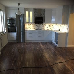 Albert Brzezin - Nowoczesna, jasna kuchnia z drewnianą podłogą, białymi szafkami i mozaiką nad blatem, widok na całe pomieszczenie.