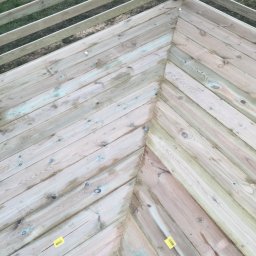 Albert Brzezin - Drewniany narożnik tarasu z balustradą, widok z góry, deski ułożone w jodełkę, widoczne ślady wilgoci i żółte naklejki.