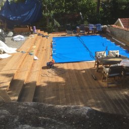 Albert Brzezin - Nowy drewniany taras z widocznymi schodami, częściowo zakryty niebieską plandeką, z meblami ogrodowymi i zabawką, otoczony zielenią.