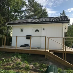 Biały domek narzędziowy z drewnianym tarasem i czarną skrzynką pocztową, otoczony zielenią i drzewami. Widoczna rolka siatki ogrodzeniowej.