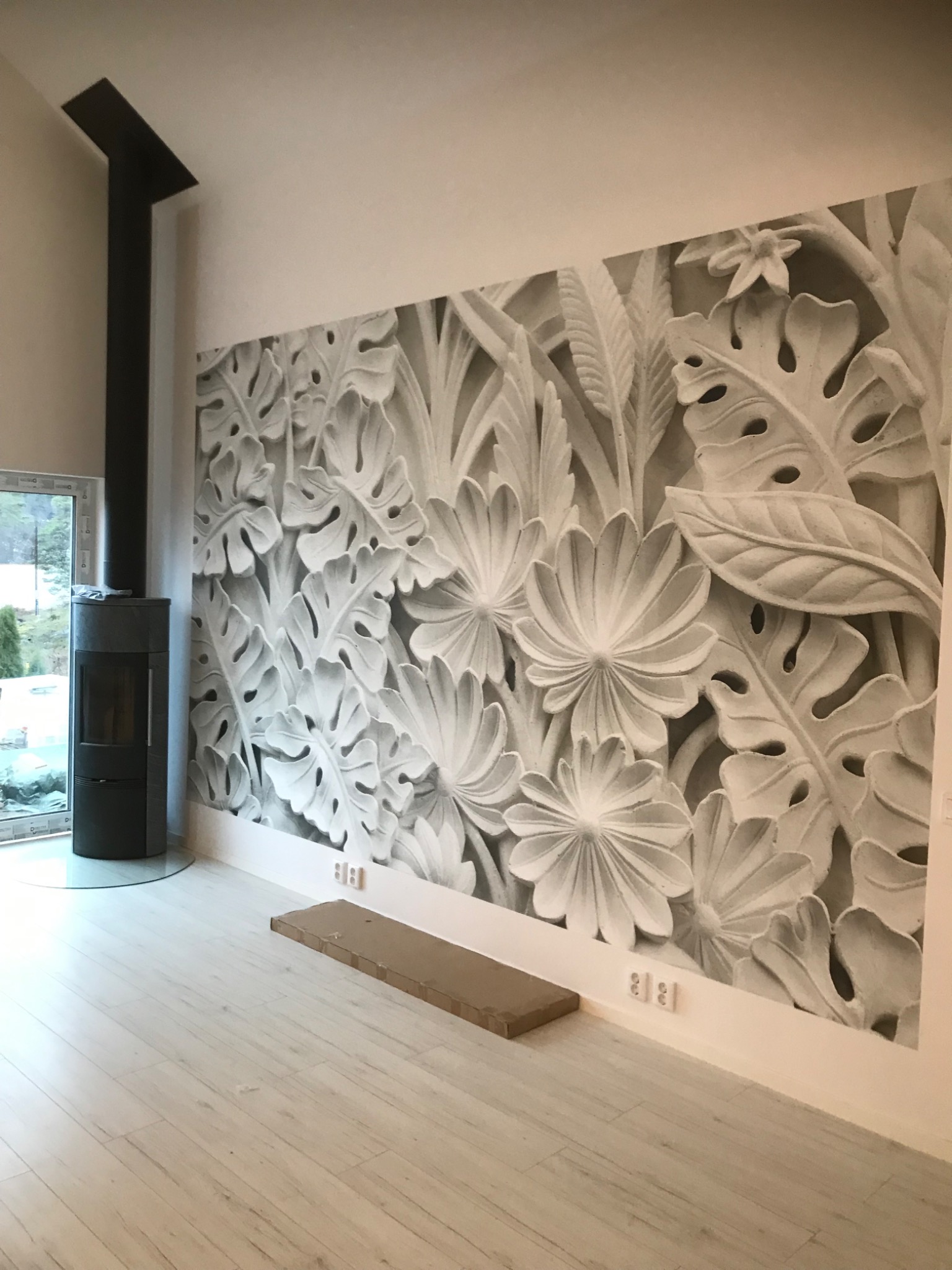 Salon z nowoczesnym piecem wolnostojącym i fototapetą z motywem liści monstery i kwiatów w odcieniach szarości, drewniana podłoga, widok na zewnątrz przez okno.