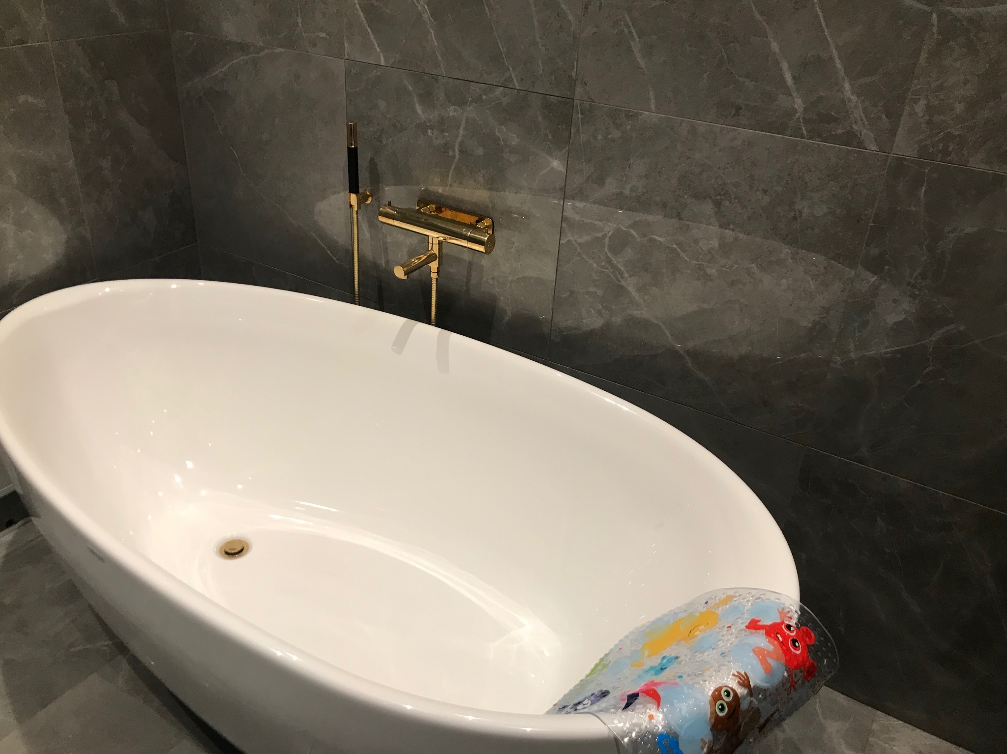 Nowoczesna, wolnostojąca wanna owalna z armaturą w kolorze złotym i szarymi płytkami imitującymi marmur na ścianie, z widoczną nakładką antypoślizgową z motywami zwierzęcymi.