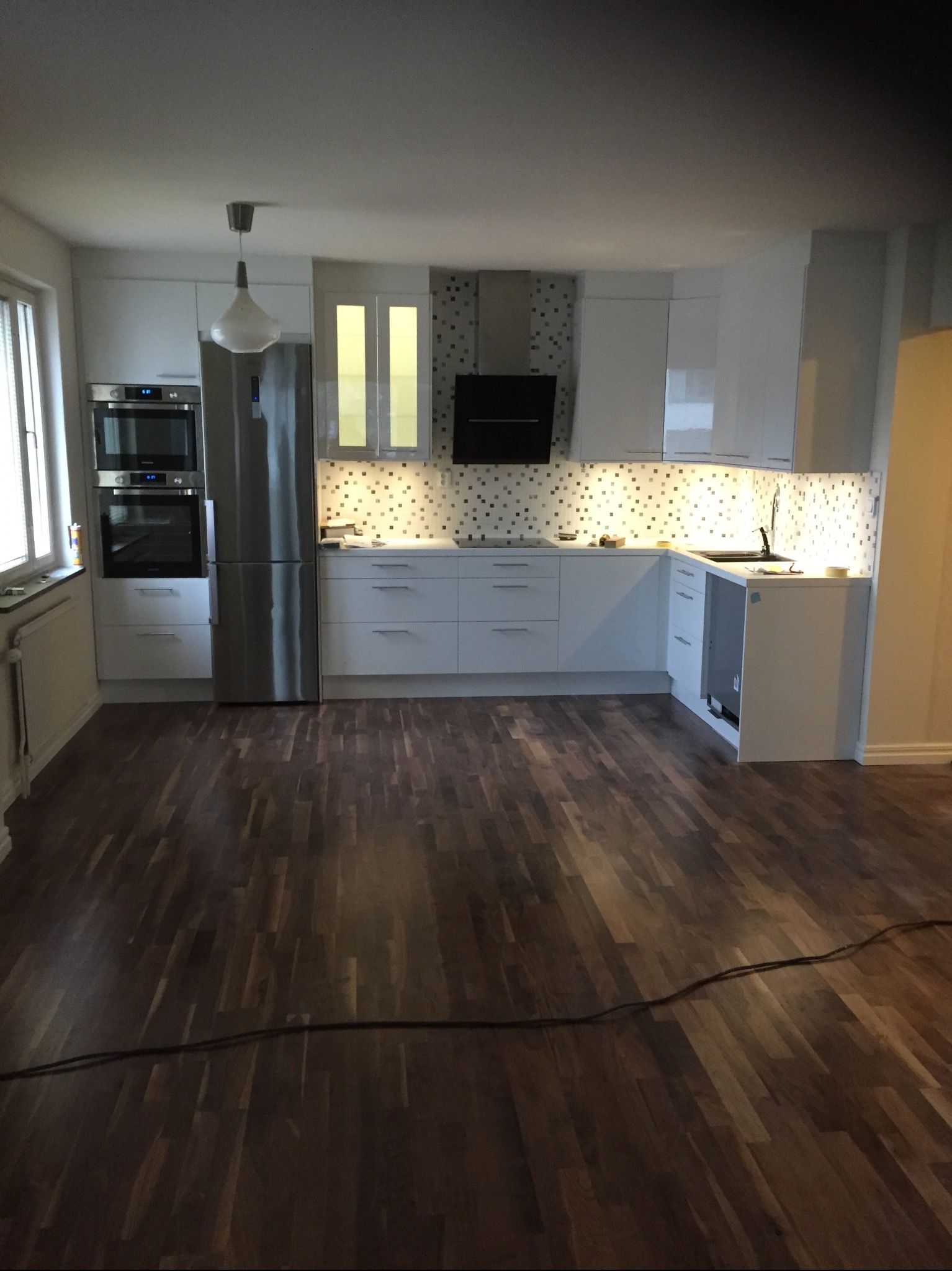 Nowoczesna, jasna kuchnia z drewnianą podłogą, białymi szafkami i mozaiką nad blatem, widok na całe pomieszczenie.