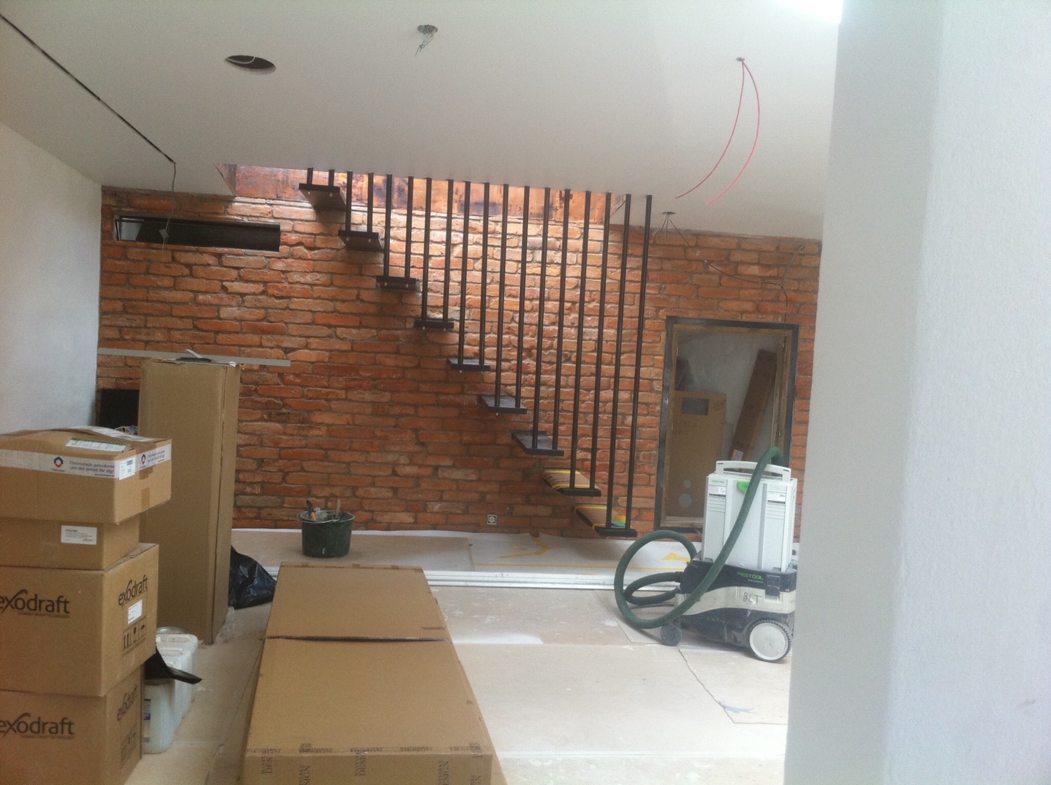 Wnętrze w trakcie prac wykończeniowych: surowa ceglana ściana z metalowymi schodami, widoczne kartony, narzędzia i odkurzacz przemysłowy Festool.