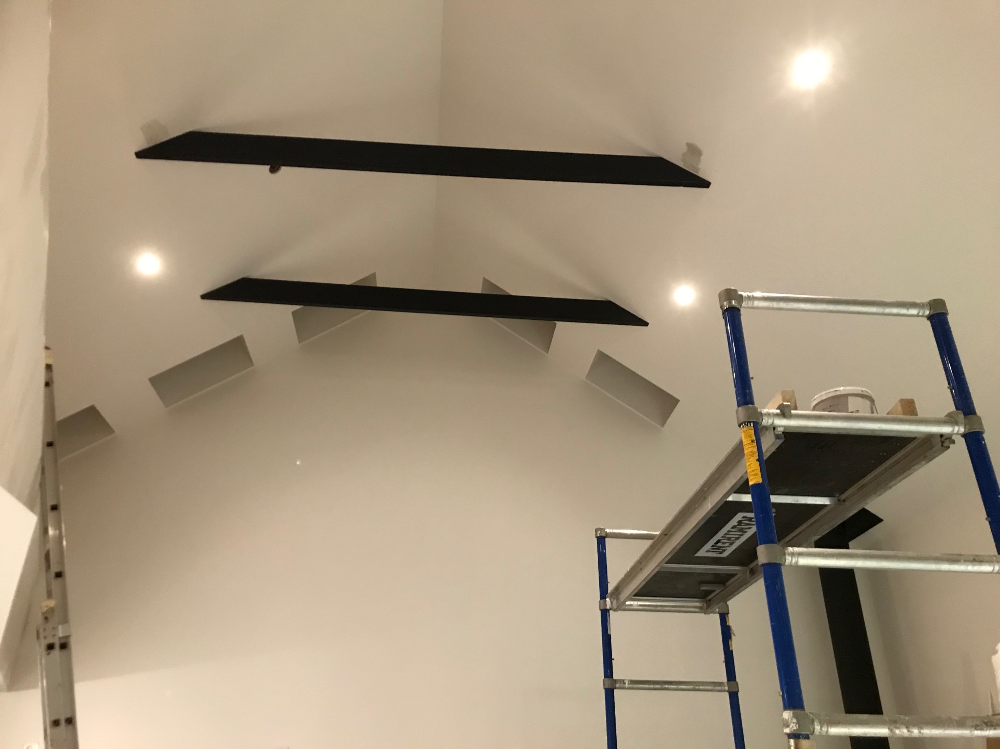 Biały sufit skośny z zamontowanymi czarnymi profilami i wnękami oświetleniowymi, widoczny fragment rusztowania malarskiego z wiadrem farby.