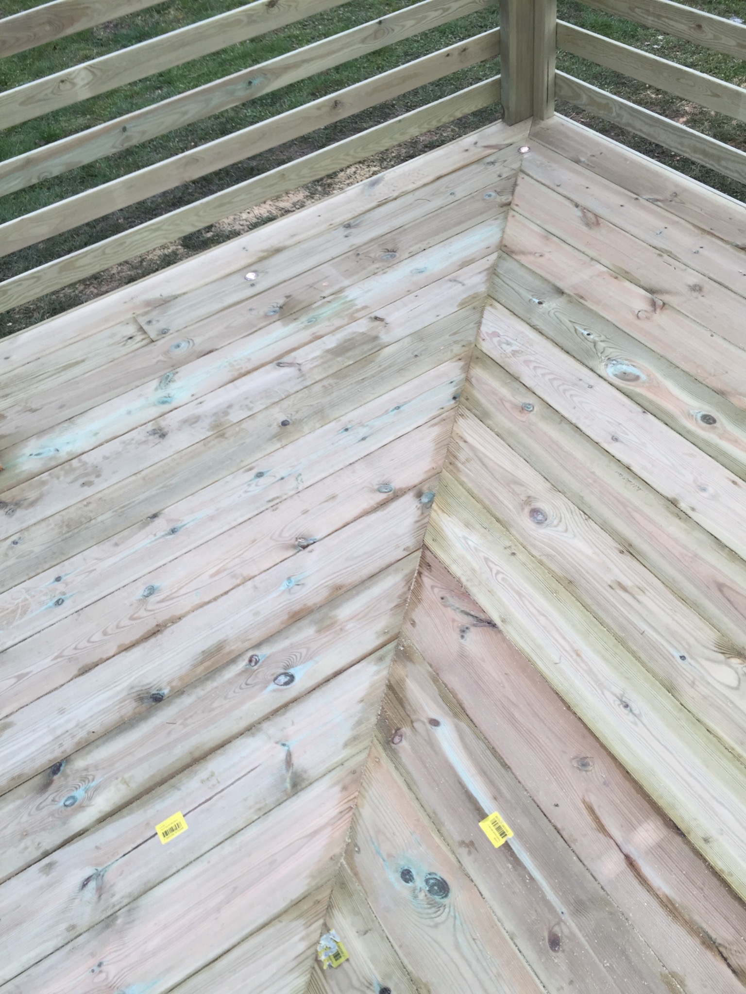 Drewniany narożnik tarasu z balustradą, widok z góry, deski ułożone w jodełkę, widoczne ślady wilgoci i żółte naklejki.