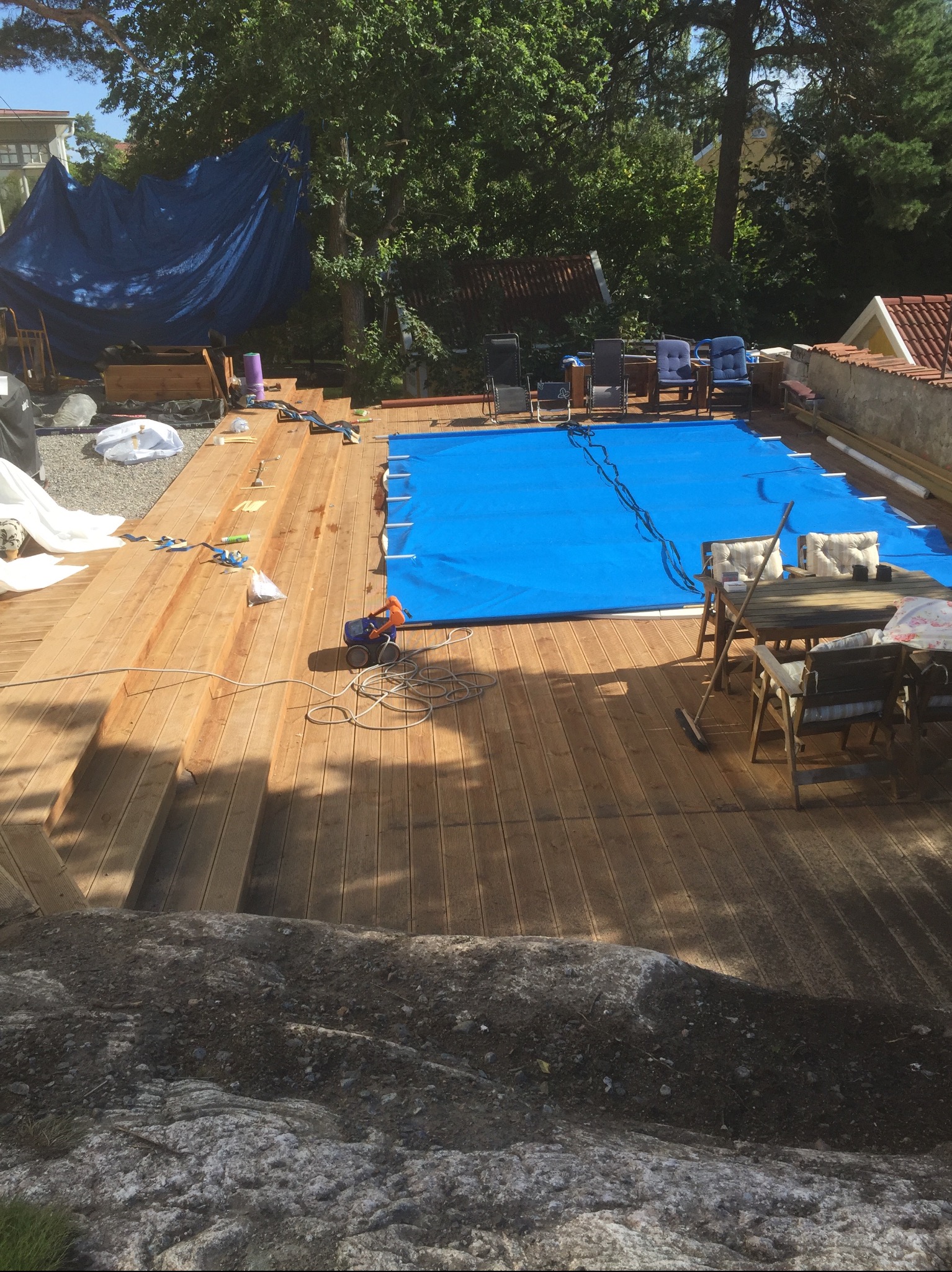 Nowy drewniany taras z widocznymi schodami, częściowo zakryty niebieską plandeką, z meblami ogrodowymi i zabawką, otoczony zielenią.