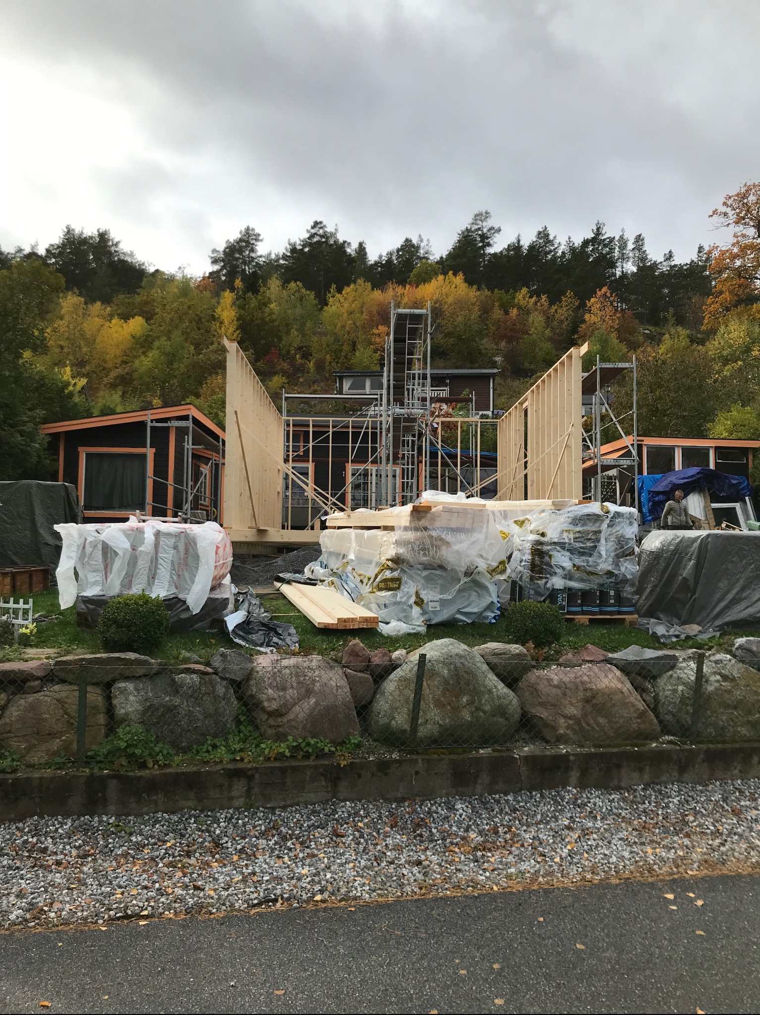 Budowa szkieletowej konstrukcji domu z widocznymi elementami drewnianymi, rusztowaniami i materiałami budowlanymi na tle jesiennego krajobrazu.