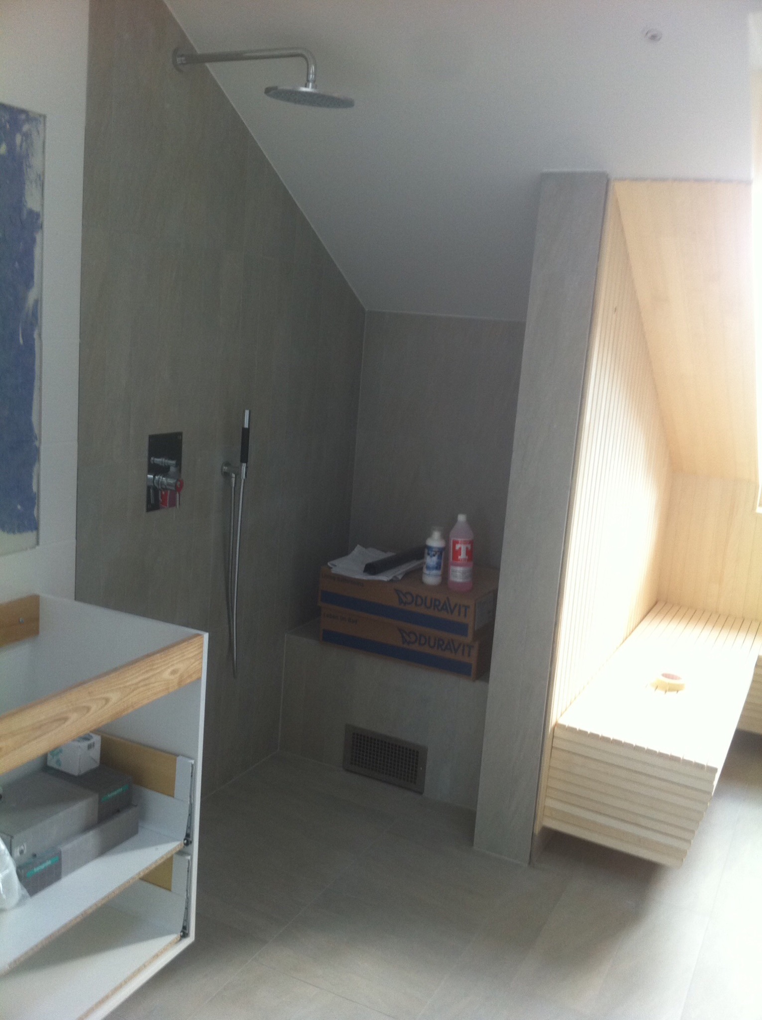 Nowoczesna łazienka z kabiną prysznicową wyłożoną płytkami w odcieniach szarości i drewnianą sauną. Widoczne akcesoria łazienkowe i fragment mebli w trakcie montażu.