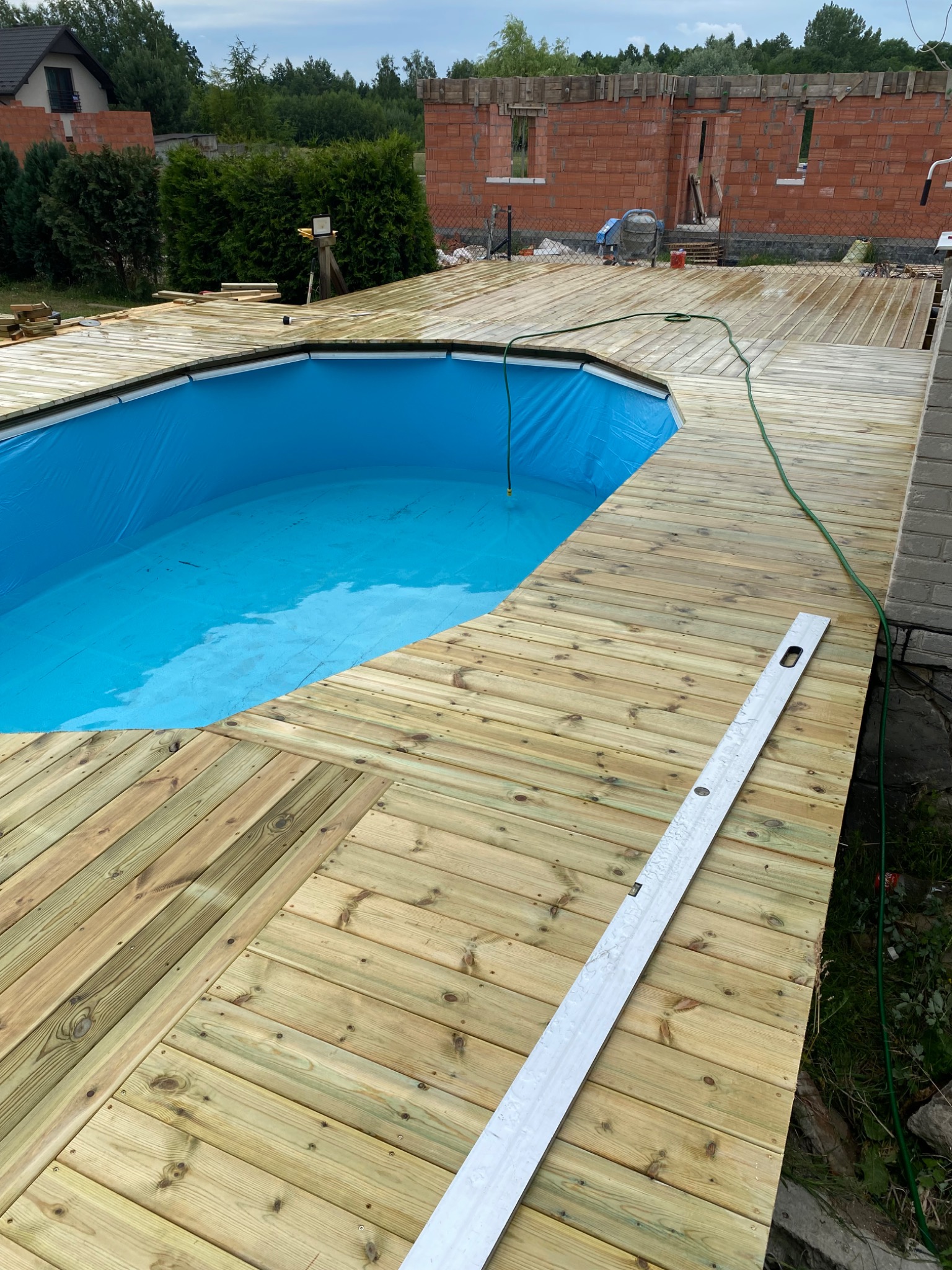 Drewniany taras wokół basenu z niebieską folią, poziomica na deskach, w tle budowa domu z czerwonej cegły i zielone krzewy. Elementy mokre.
