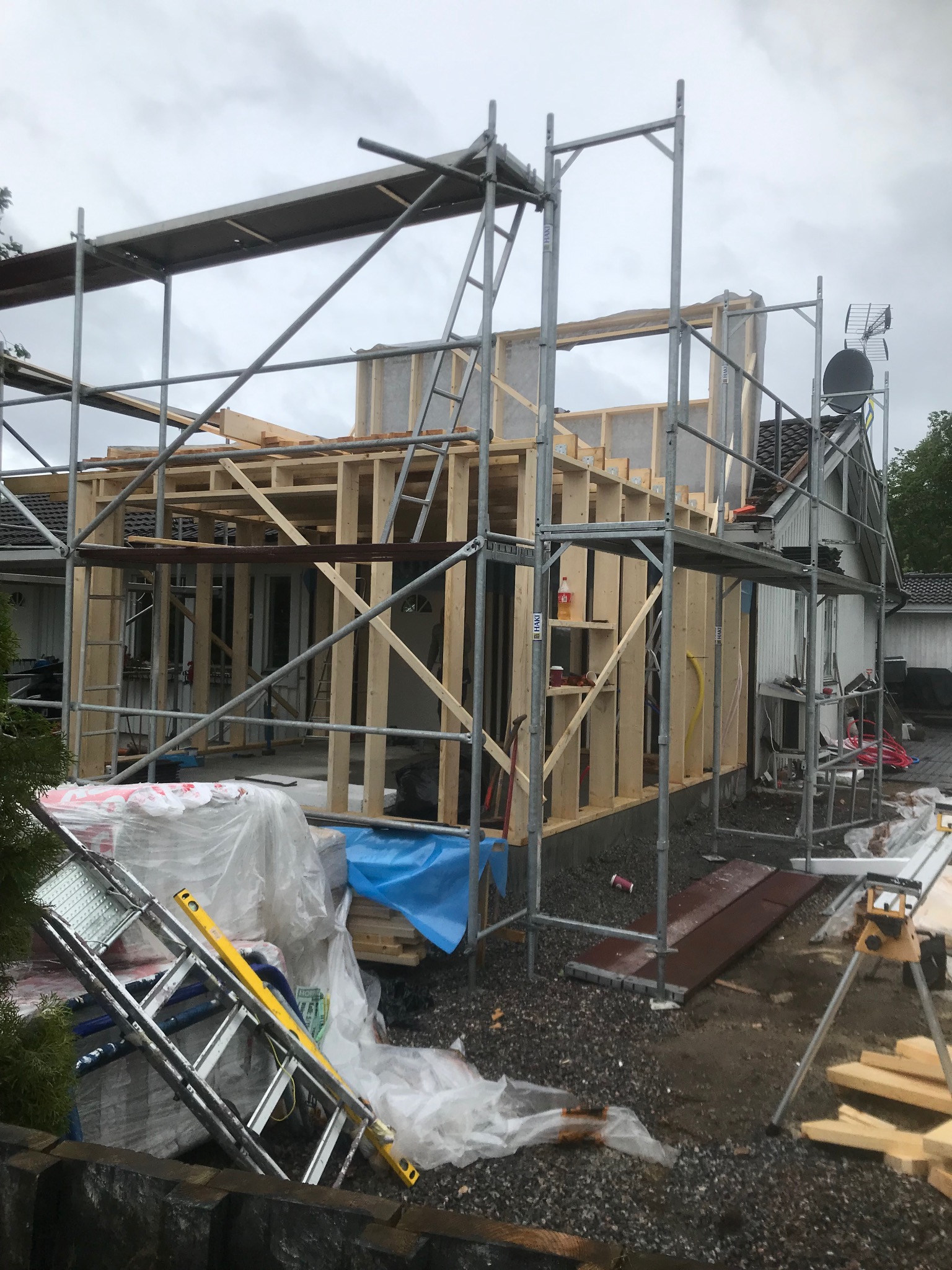 Budowa domu w trakcie realizacji: drewniana konstrukcja szkieletowa, rusztowania, materiały budowlane. Widoczny fragment istniejącego budynku.