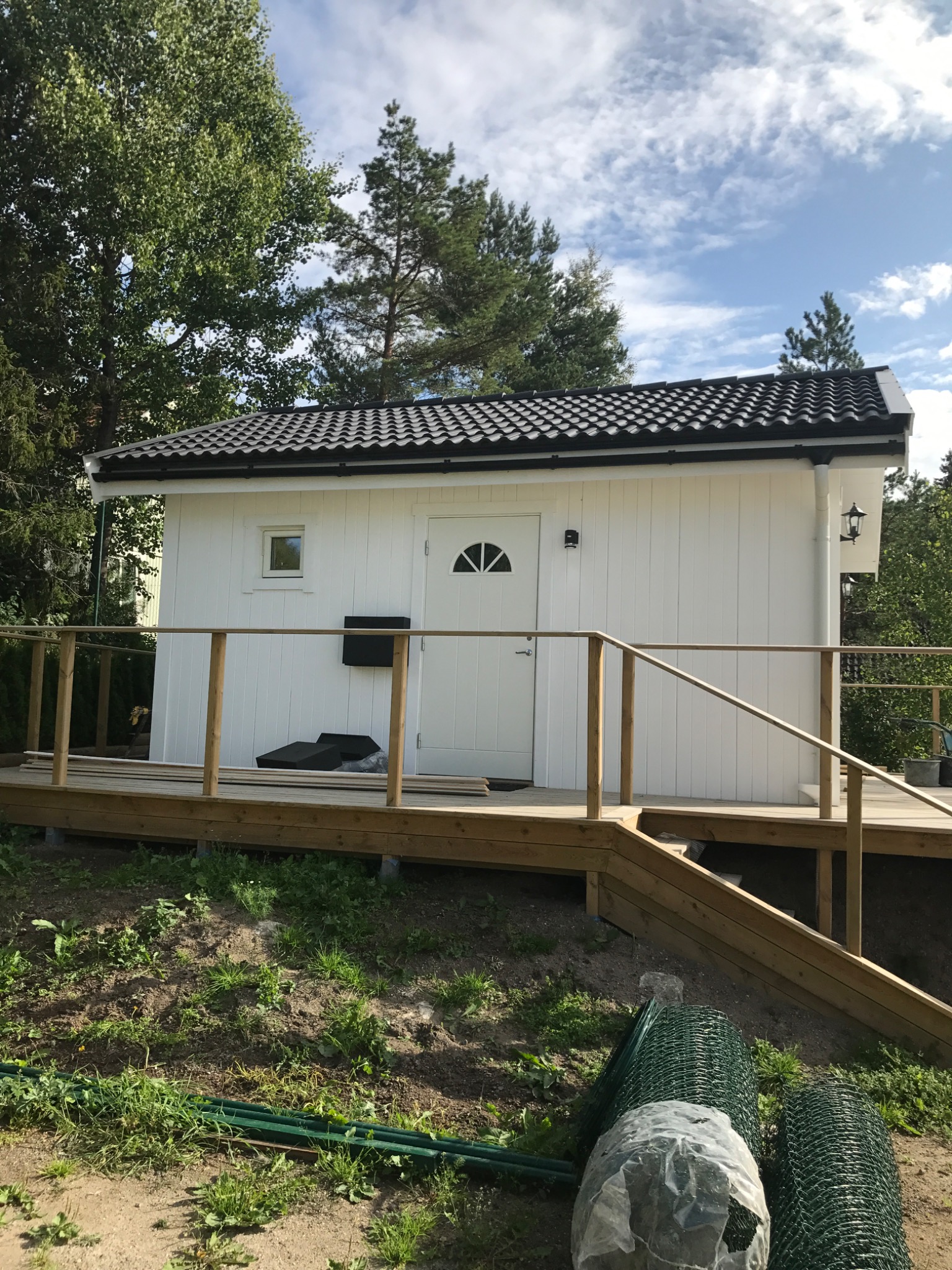 Biały domek narzędziowy z drewnianym tarasem i czarną skrzynką pocztową, otoczony zielenią i drzewami. Widoczna rolka siatki ogrodzeniowej.