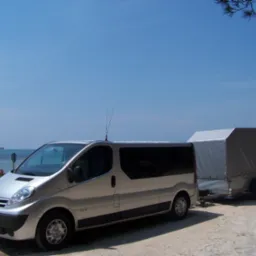 Srebrny bus Nissan z przyczepą na tle błękitnego nieba i morza, antena na dachu, widoczne drzewa i słup wysokiego napięcia w oddali.