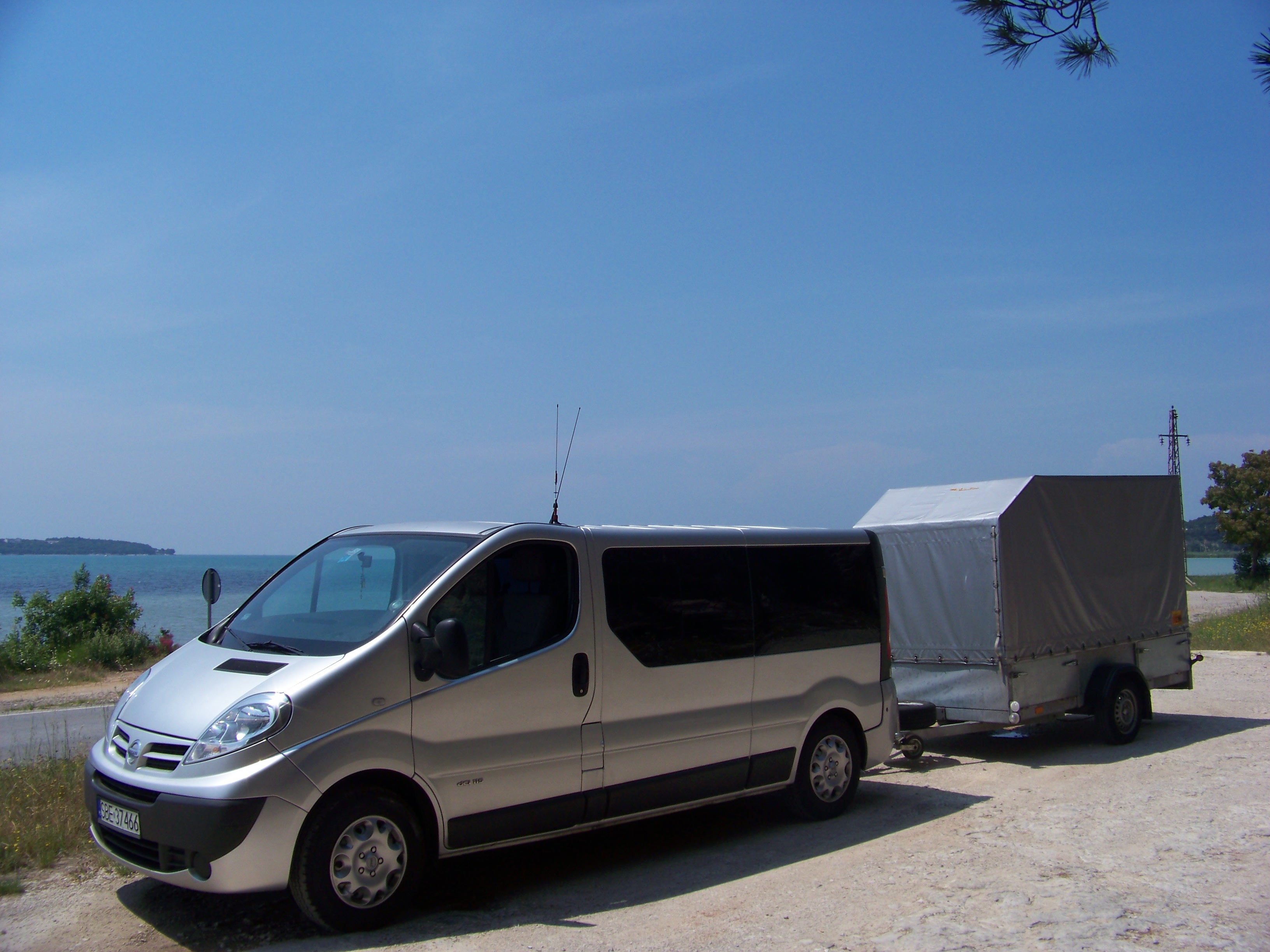 Srebrny bus Nissan z przyczepą na tle błękitnego nieba i morza, antena na dachu, widoczne drzewa i słup wysokiego napięcia w oddali.