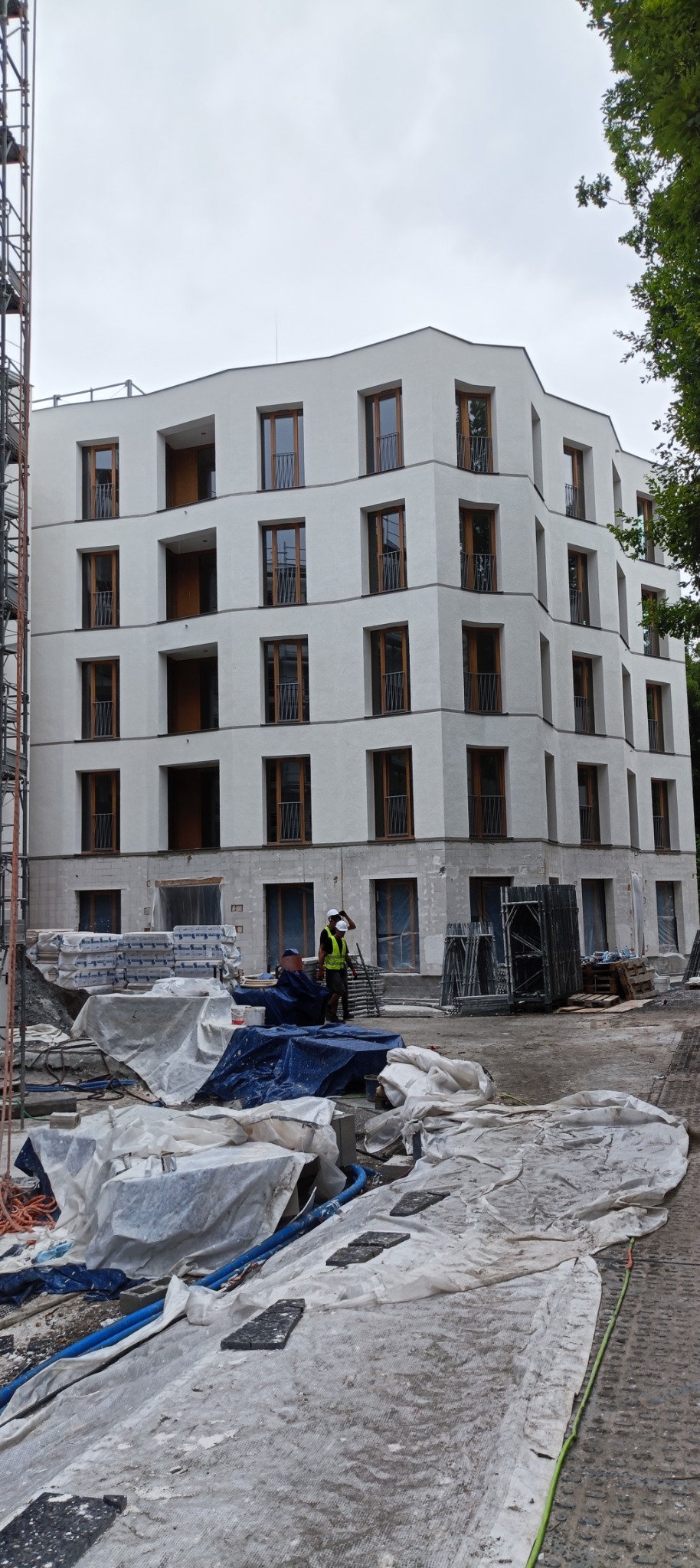 Wielopiętrowy budynek mieszkalny z białymi elewacjami i drewnianymi oknami w trakcie budowy, widoczne materiały budowlane i pracownicy w kaskach i kamizelkach odblaskowych.